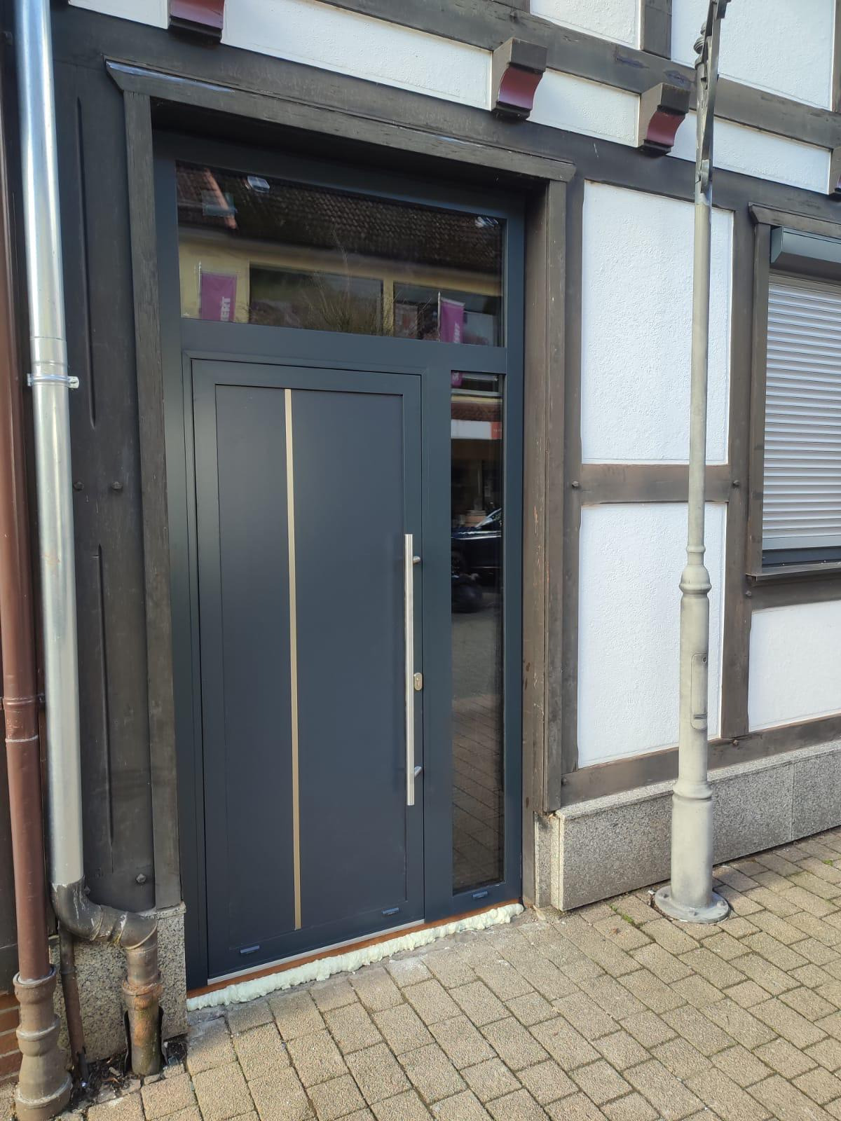Grafitowe drzwi wejściowe z pionowym, złotym akcentem i długim, srebrnym uchwytem, zamontowane w budynku o konstrukcji szachulcowej, widoczne ślady montażu w postaci pianki uszczelniającej.