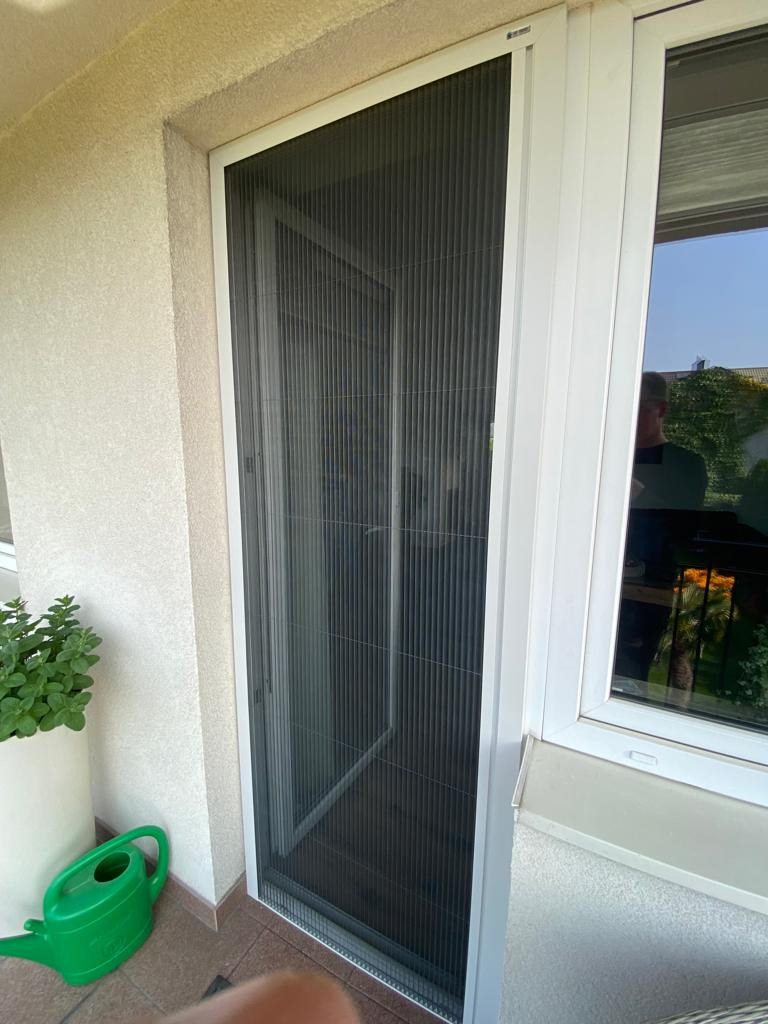 Zewnętrzna, plisowana moskitiera w białej ramie, zamontowana przy oknie balkonowym, widok z zewnątrz, obok doniczka z zieloną rośliną i zielona konewka.