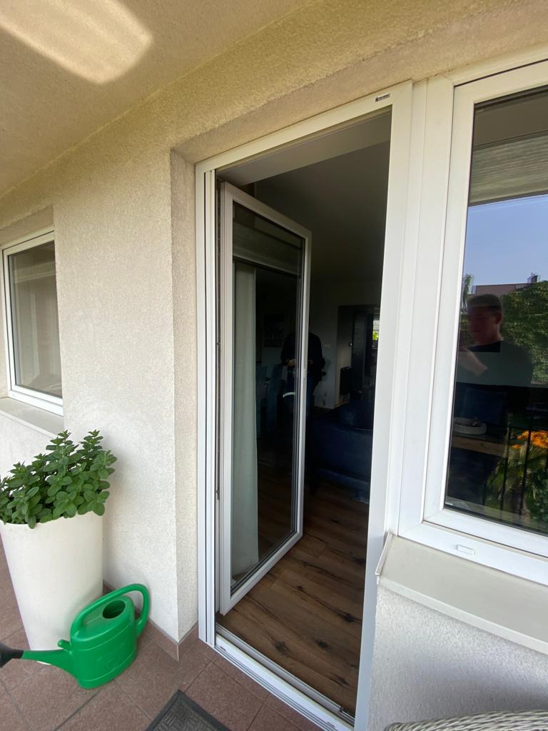 Otwarta, biała, przesuwna konstrukcja okienno-drzwiowa z widokiem na balkon z doniczką i konewką, widoczny fragment wnętrza z drewnianą podłogą.
