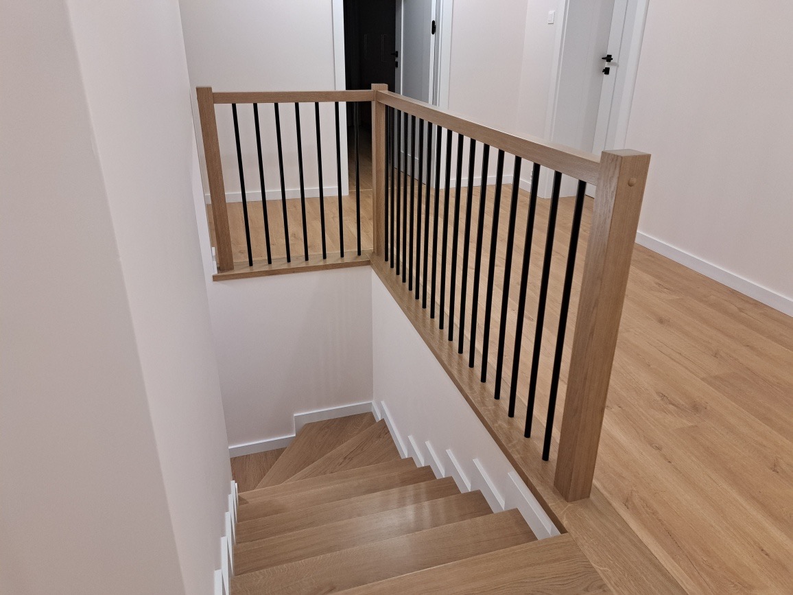 Jasne, drewniane schody z czarnymi, metalowymi prętami balustrady i drewnianą poręczą, widok z góry na dół w nowoczesnym wnętrzu.