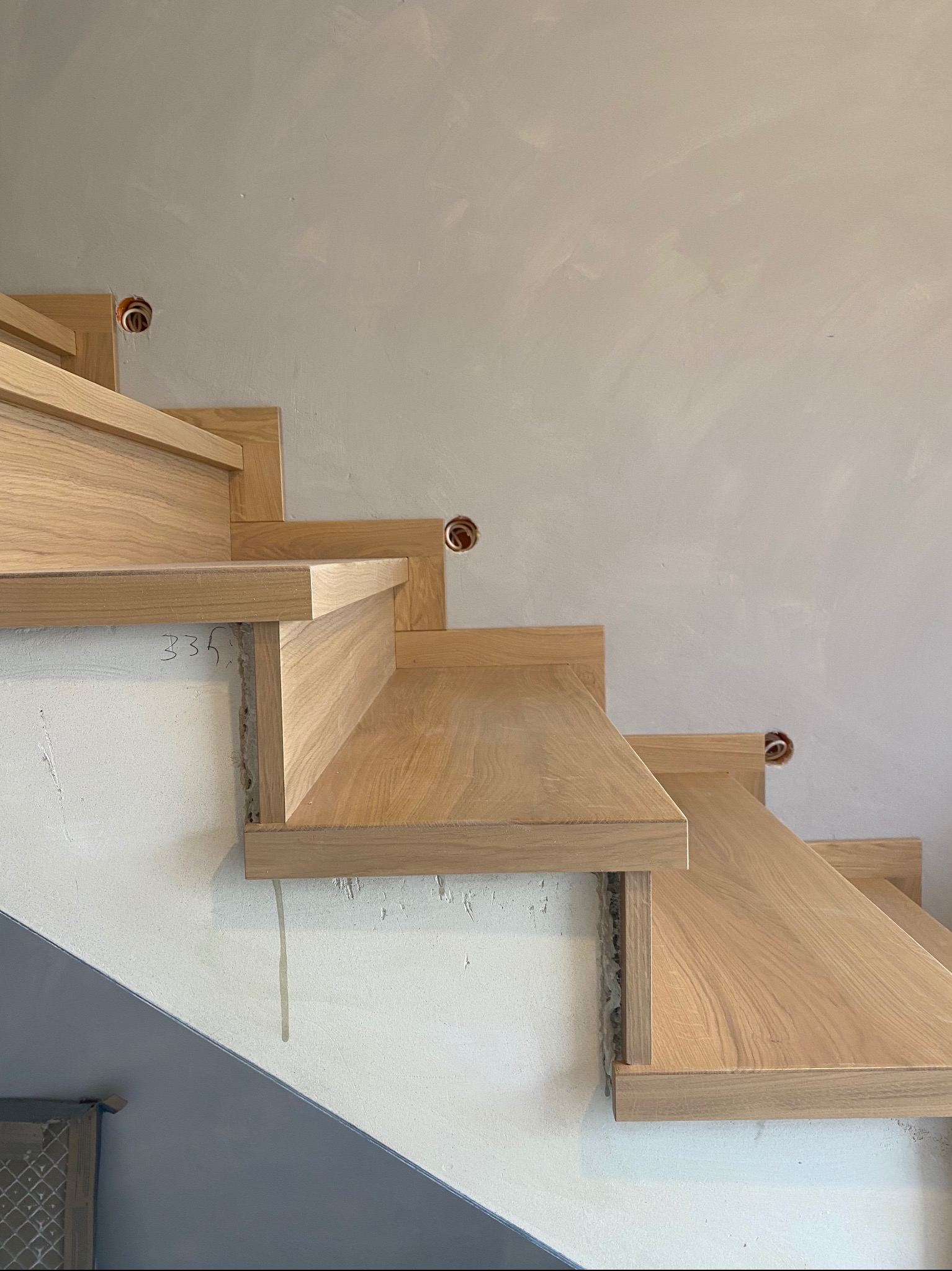 Surowe, dębowe stopnie schodowe w trakcie montażu, widoczne niedoskonałości ściany i otwory na kable.