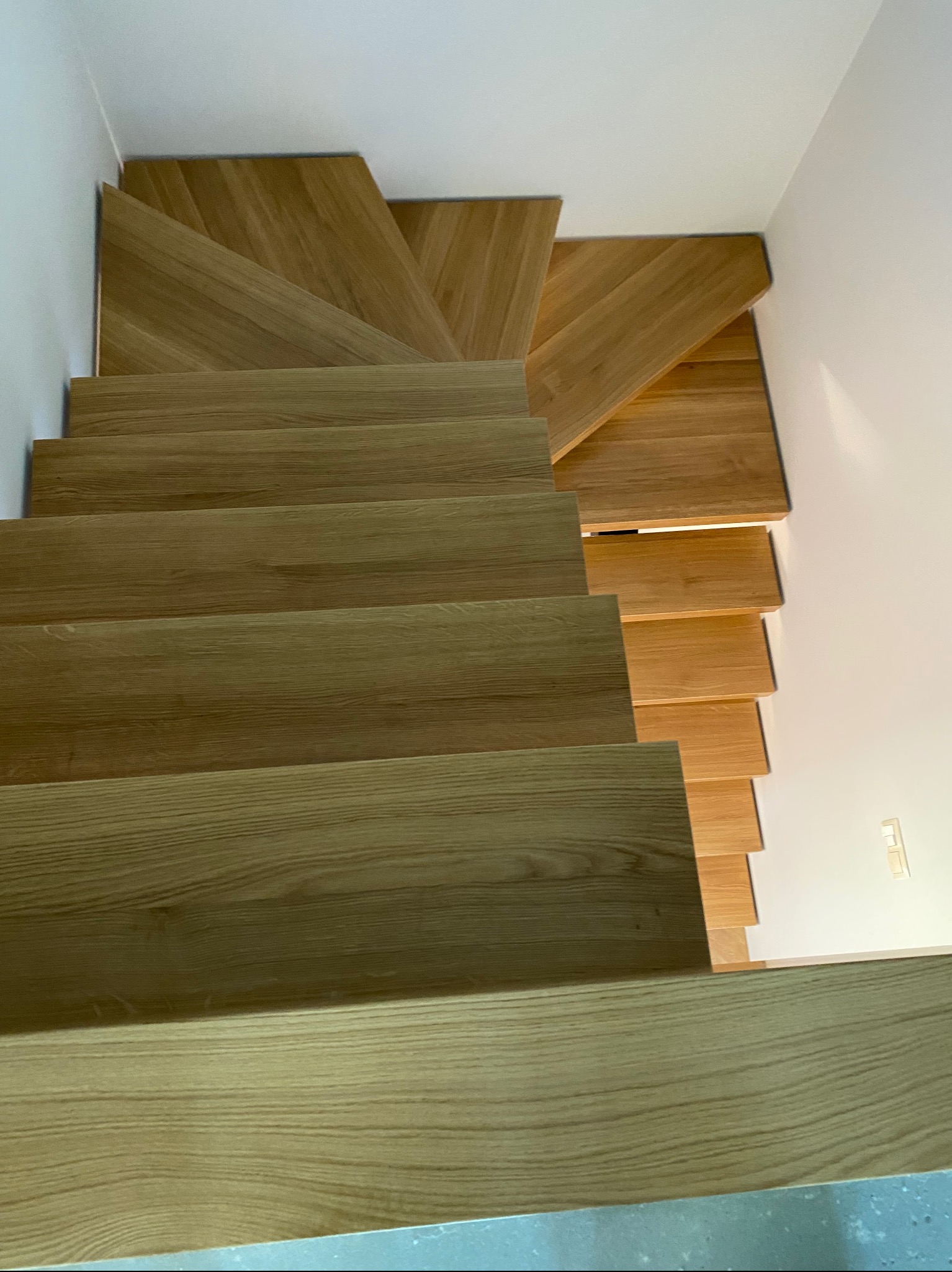 Widok z góry na nowoczesne schody z jasnego drewna dębowego, z widocznym usłojeniem, zamontowane przy białej ścianie.