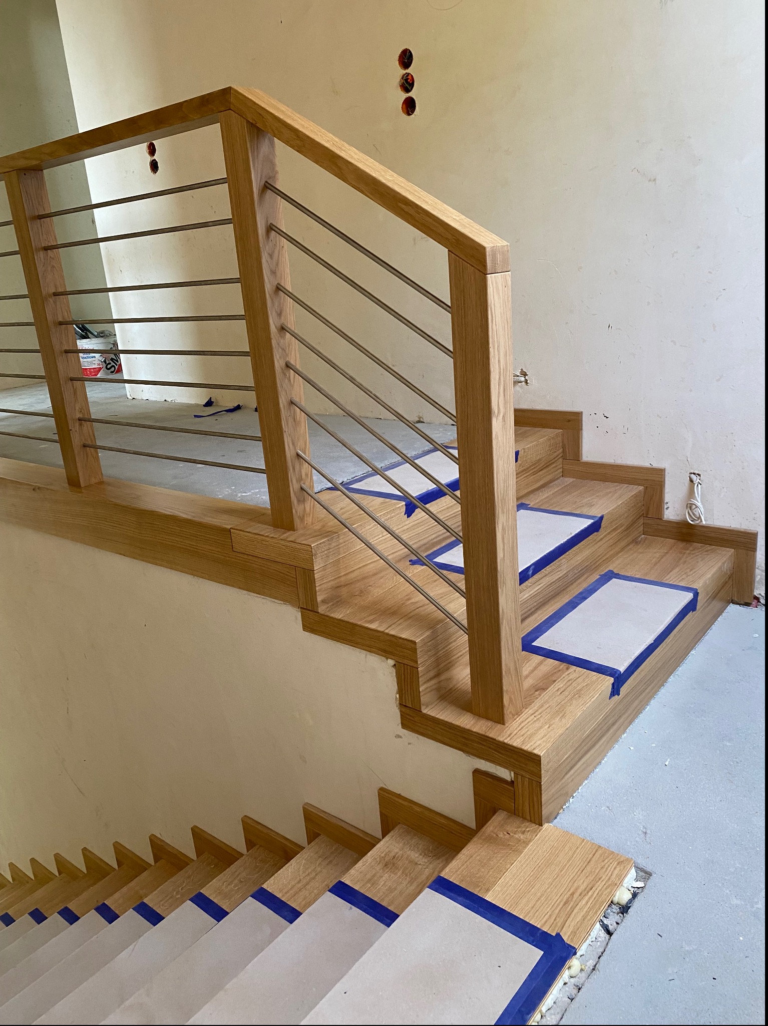 Montaż nowoczesnych schodów z drewnianymi stopniami zabezpieczonymi kartonem i niebieską taśmą, z metalową balustradą, w surowym wnętrzu.
