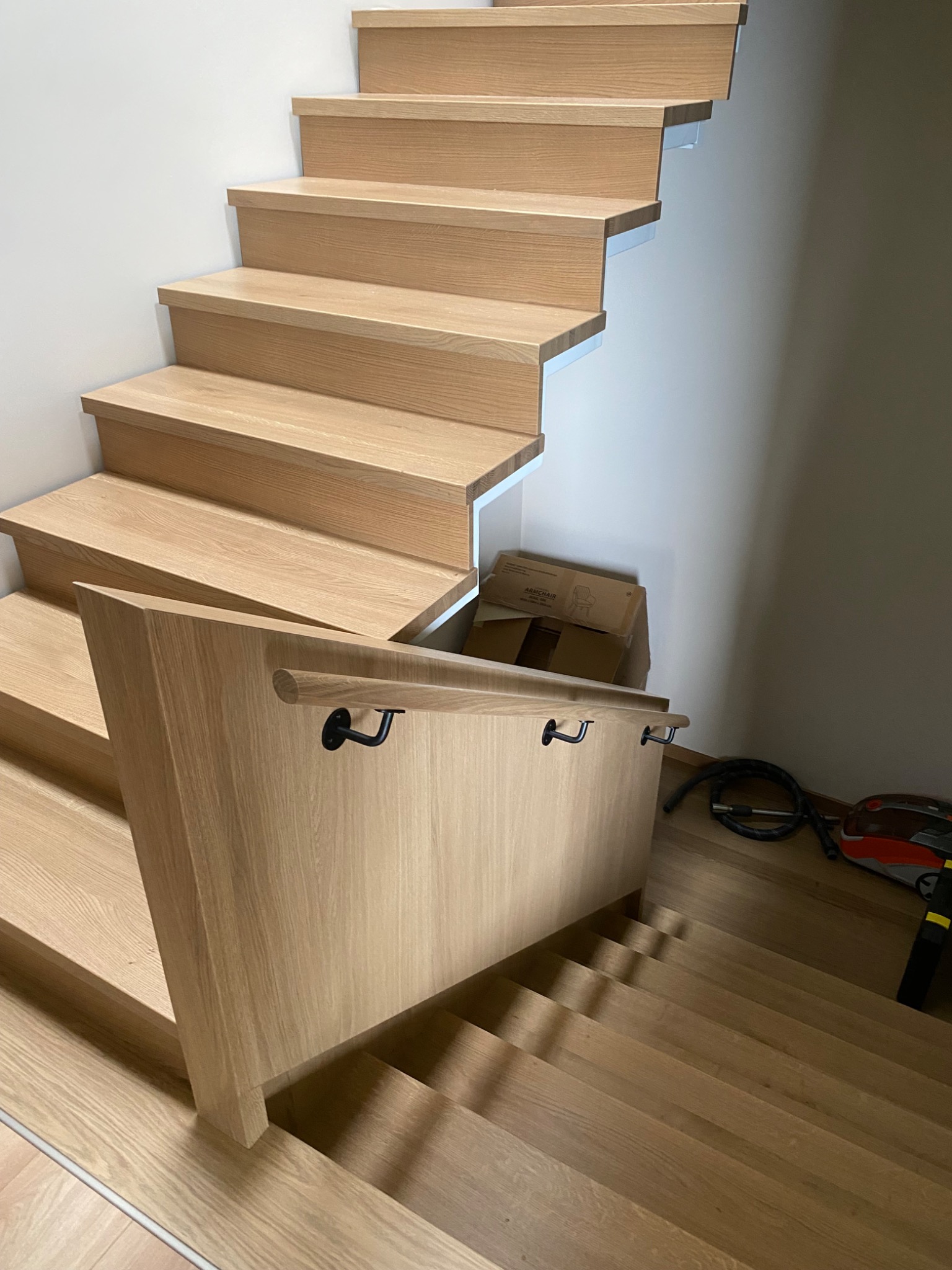 Nowoczesne, jasne schody z drewna z czarnymi uchwytami poręczy, widoczne z góry, z elementami montażowymi i kartonem w tle. Minimalistyczny design.