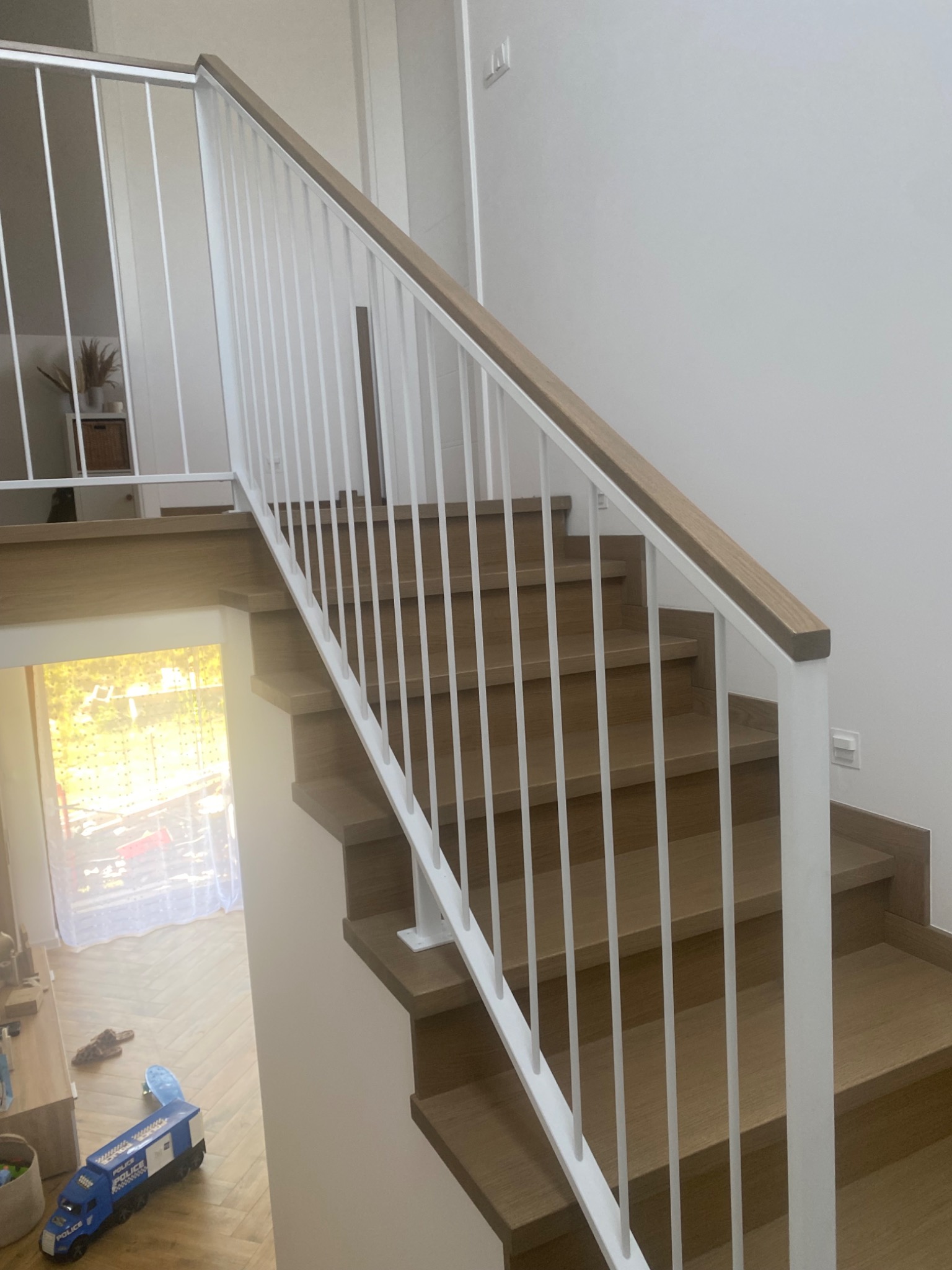 Nowoczesne, drewniane schody z białą balustradą. Jasne drewno stopni kontrastuje z białymi elementami. W tle widoczny pokój dziecięcy i światło wpadające przez okno.