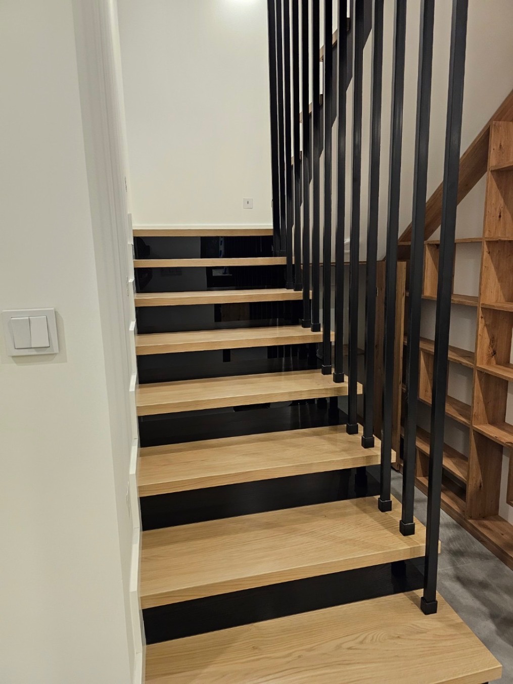 Nowoczesne schody z jasnego drewna z czarnymi podstopnicami i minimalistyczną, metalową balustradą. W tle regał z drewnianymi półkami.