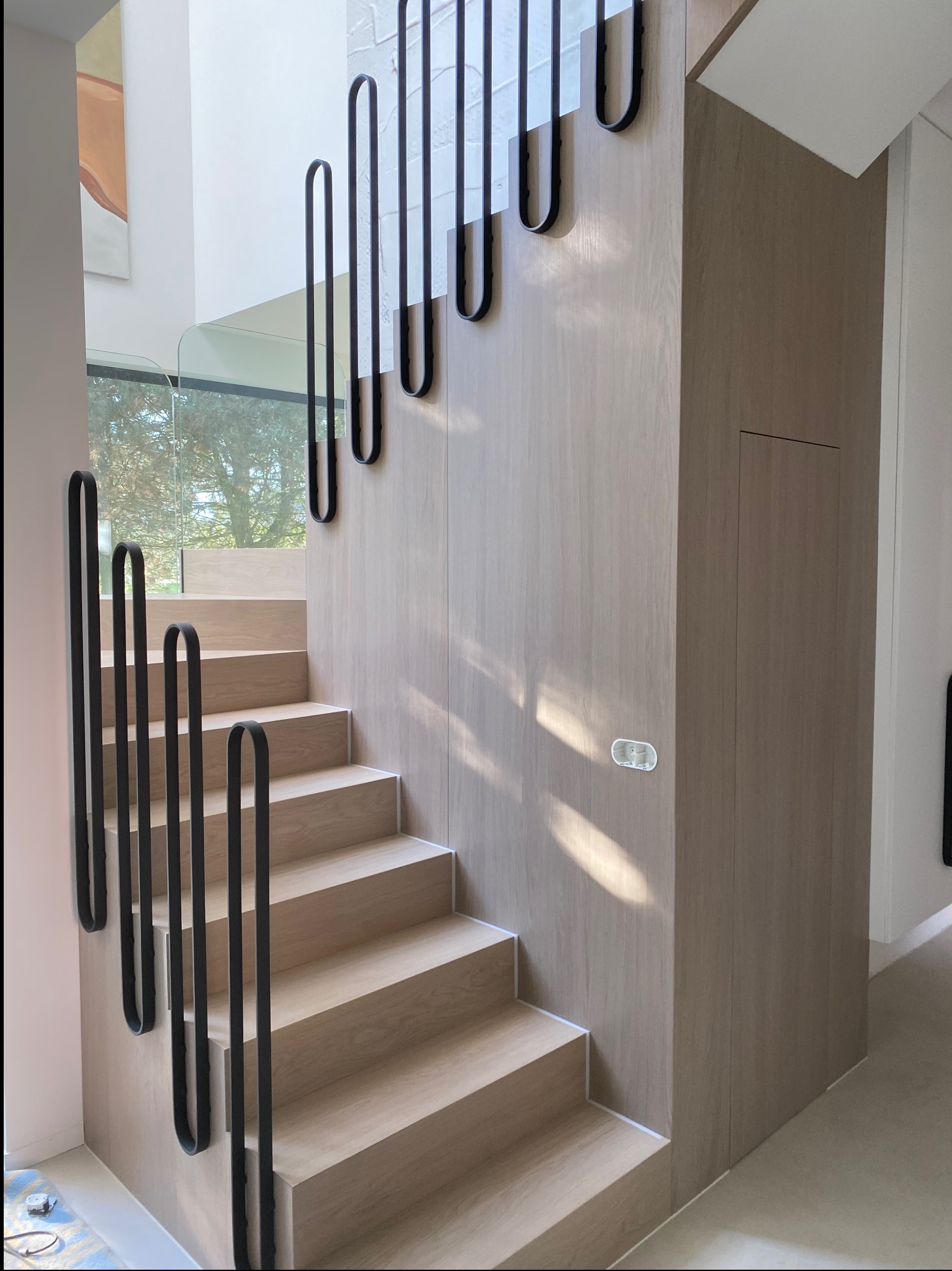 Nowoczesne schody z jasnego drewna z czarnymi, metalowymi poręczami o unikalnym, zaokrąglonym kształcie. Szklana balustrada na podeście. W tle widok na zieleń.