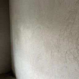 FHU Jakubiak - Świeżo nałożona, jasnoszara, strukturalna szpachla na ścianie wewnętrznej w rogu pomieszczenia, widoczne ślady narzędzi.