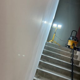 FHU Jakubiak - Gładka, świeżo wyszpachlowana ściana w trakcie schnięcia, oświetlona lampą budowlaną na trójnogu, z widocznym odkurzaczem przemysłowym na schodach.