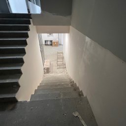 FHU Jakubiak - Klatka schodowa w surowym stanie, ściany przygotowane do malowania, widoczne ślady szpachlowania i drabina w tle.