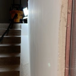 FHU Jakubiak - Świeżo pomalowana biała ściana w trakcie prac wykończeniowych, widoczny fragment schodów, żółty odkurzacz przemysłowy DeWalt i lampa oświetlająca powierzchnię.