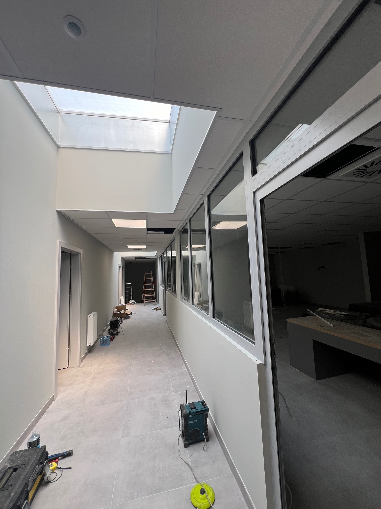Korytarz biurowy w trakcie prac wykończeniowych, widoczne narzędzia, drabina, okna z aluminiowymi ramami, świetlik w suficie i szare płytki podłogowe.
