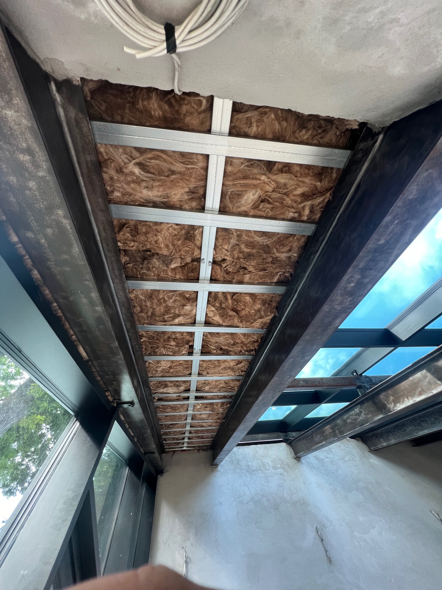 Izolacja z wełny mineralnej w metalowej konstrukcji stropu poddasza, widok z dołu, fragment okna dachowego z widocznym niebem.