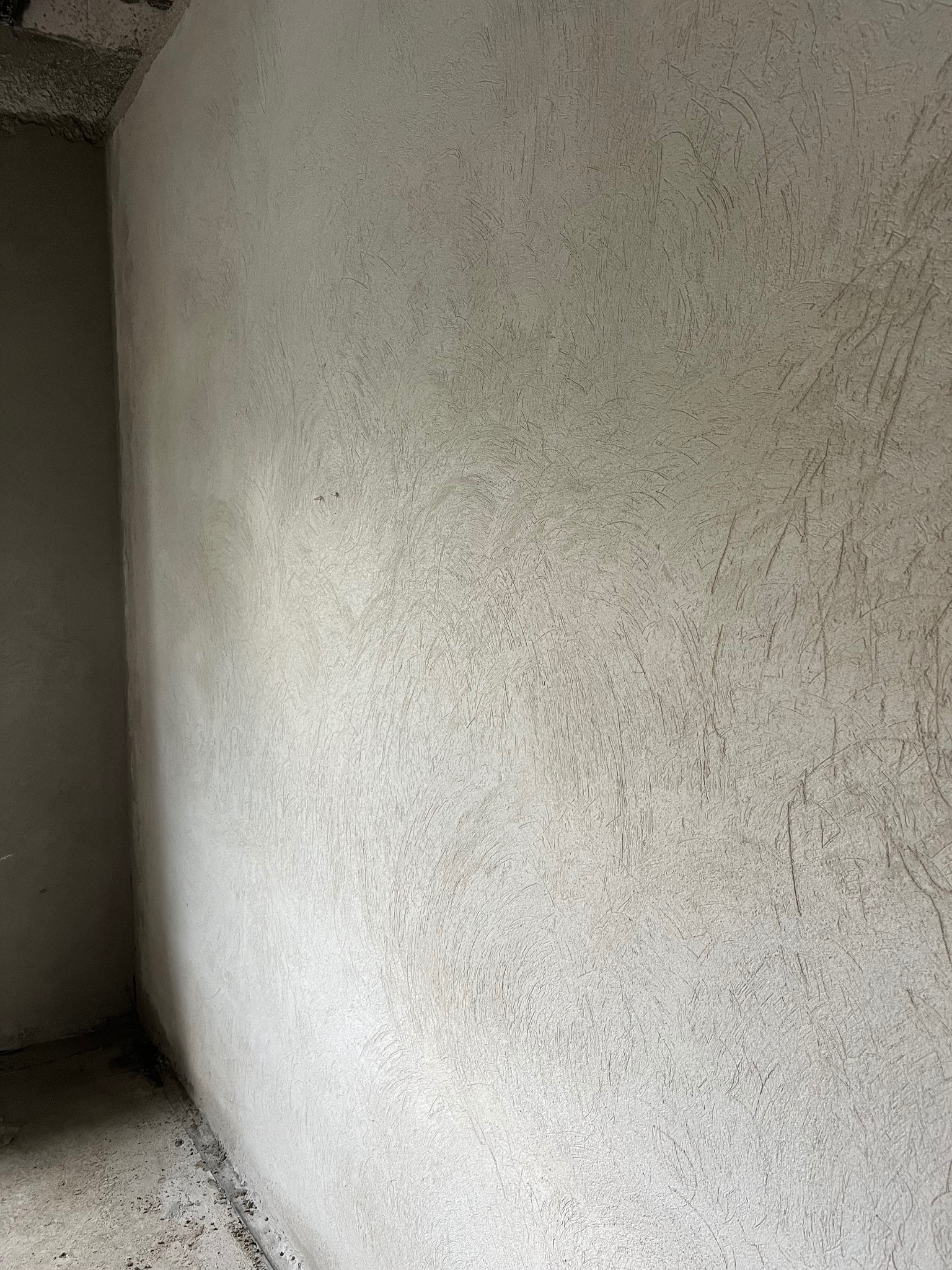 Świeżo nałożona, jasnoszara, strukturalna szpachla na ścianie wewnętrznej w rogu pomieszczenia, widoczne ślady narzędzi.