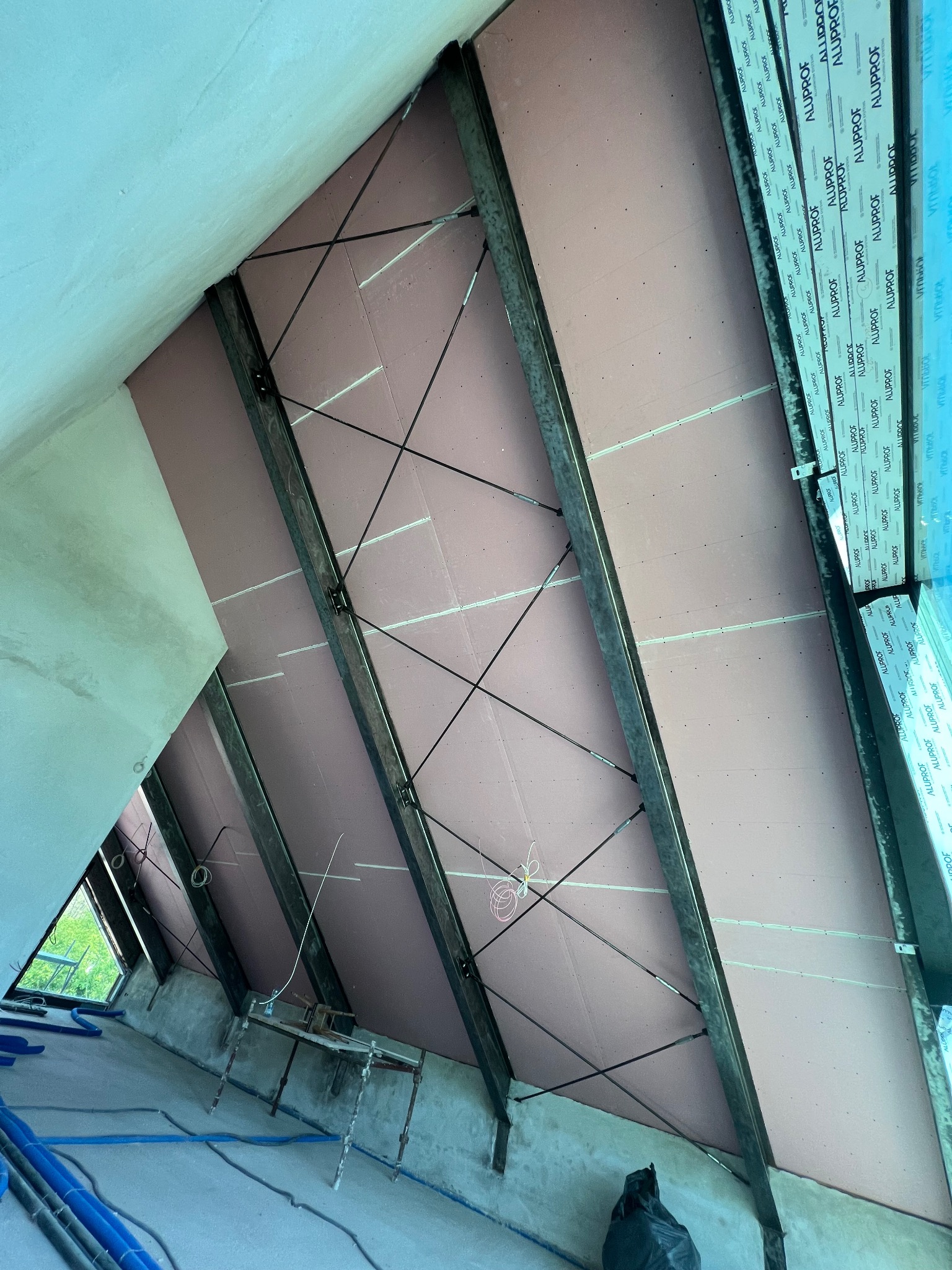 Poddasze w trakcie ocieplania, widoczne metalowe belki konstrukcyjne, wełna mineralna pokryta płytami gipsowo-kartonowymi, okno dachowe z zamontowaną stolarką okienną z widocznymi napisami...