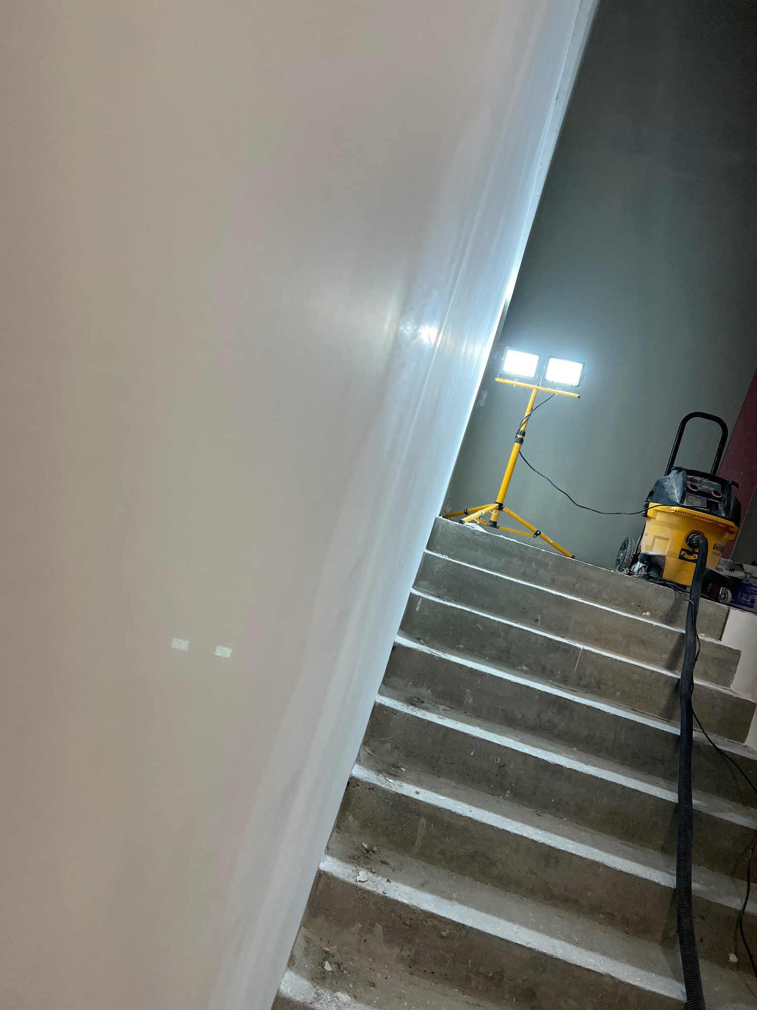 Gładka, świeżo wyszpachlowana ściana w trakcie schnięcia, oświetlona lampą budowlaną na trójnogu, z widocznym odkurzaczem przemysłowym na schodach.