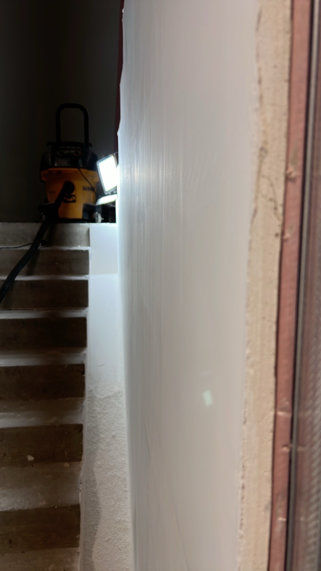 Świeżo pomalowana biała ściana w trakcie prac wykończeniowych, widoczny fragment schodów, żółty odkurzacz przemysłowy DeWalt i lampa oświetlająca powierzchnię.