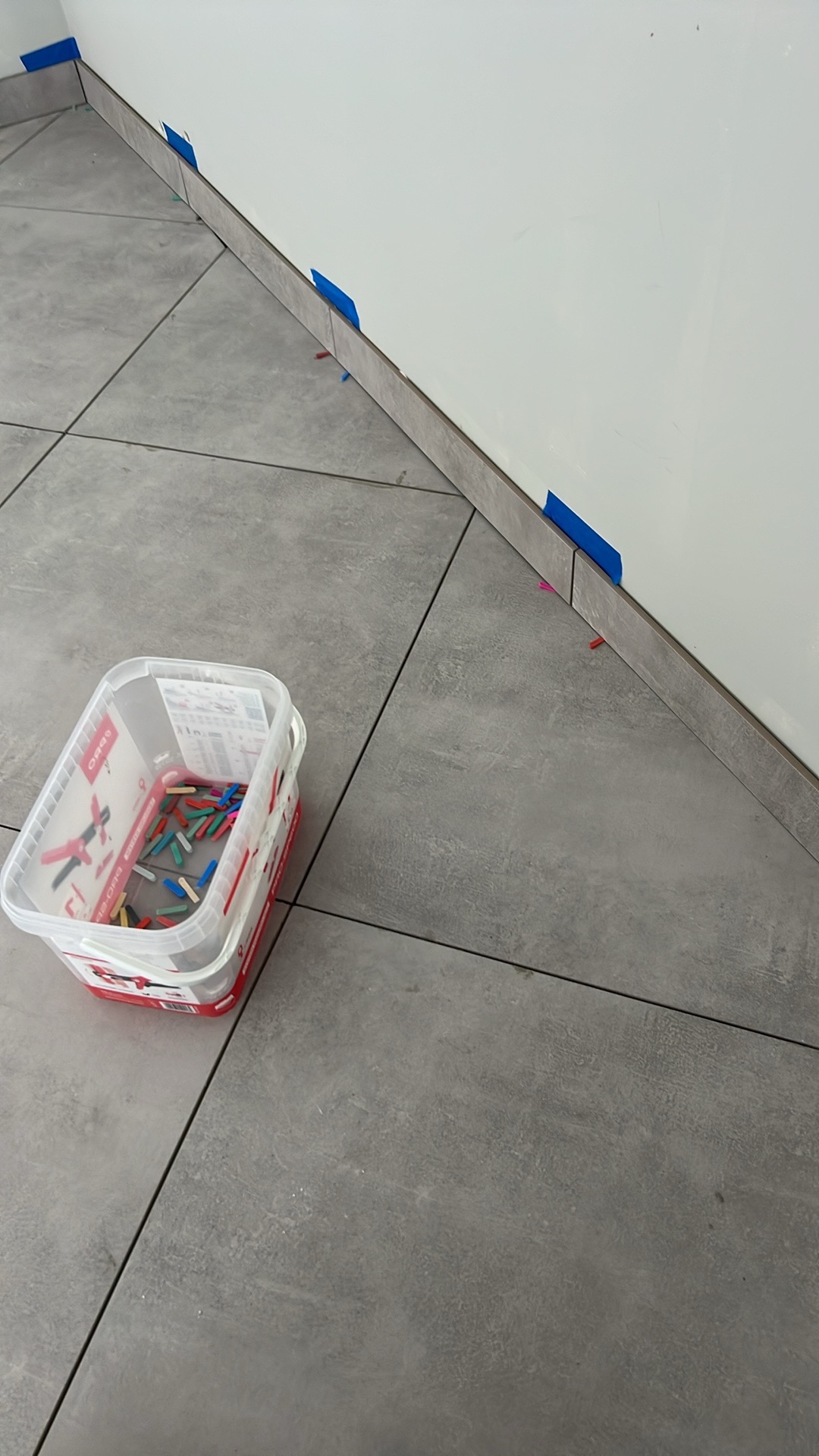 Układanie szarych płytek podłogowych wzdłuż ściany, z użyciem niebieskiej taśmy i plastikowych klinów do poziomowania, wiaderko z klinami na podłodze.
