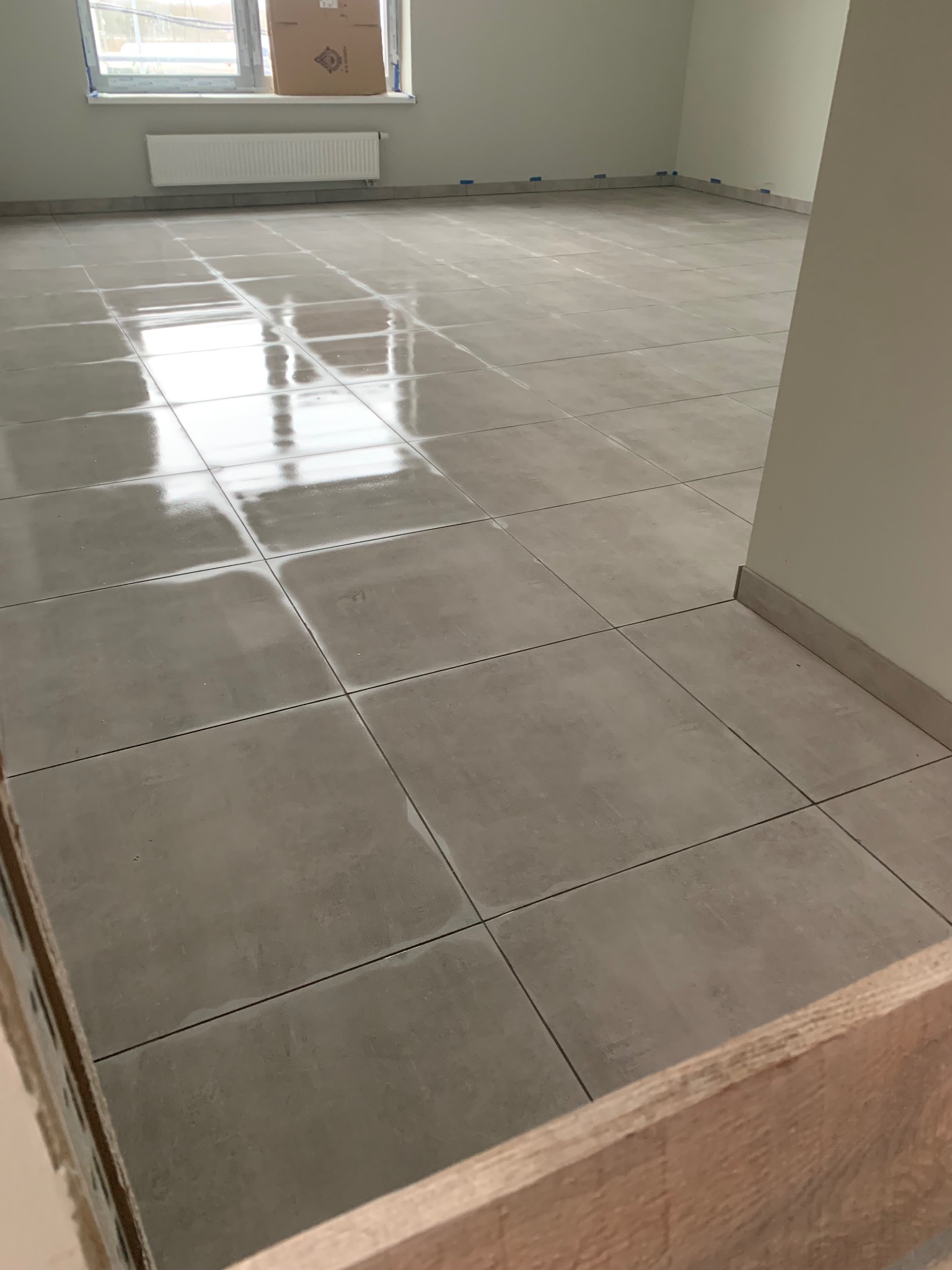 Ułożona, błyszcząca, szara podłoga z płytek ceramicznych w pustym, jasnym pomieszczeniu z grzejnikiem przy oknie. Widoczne listwy przypodłogowe i zabezpieczenia taśmą malarską.