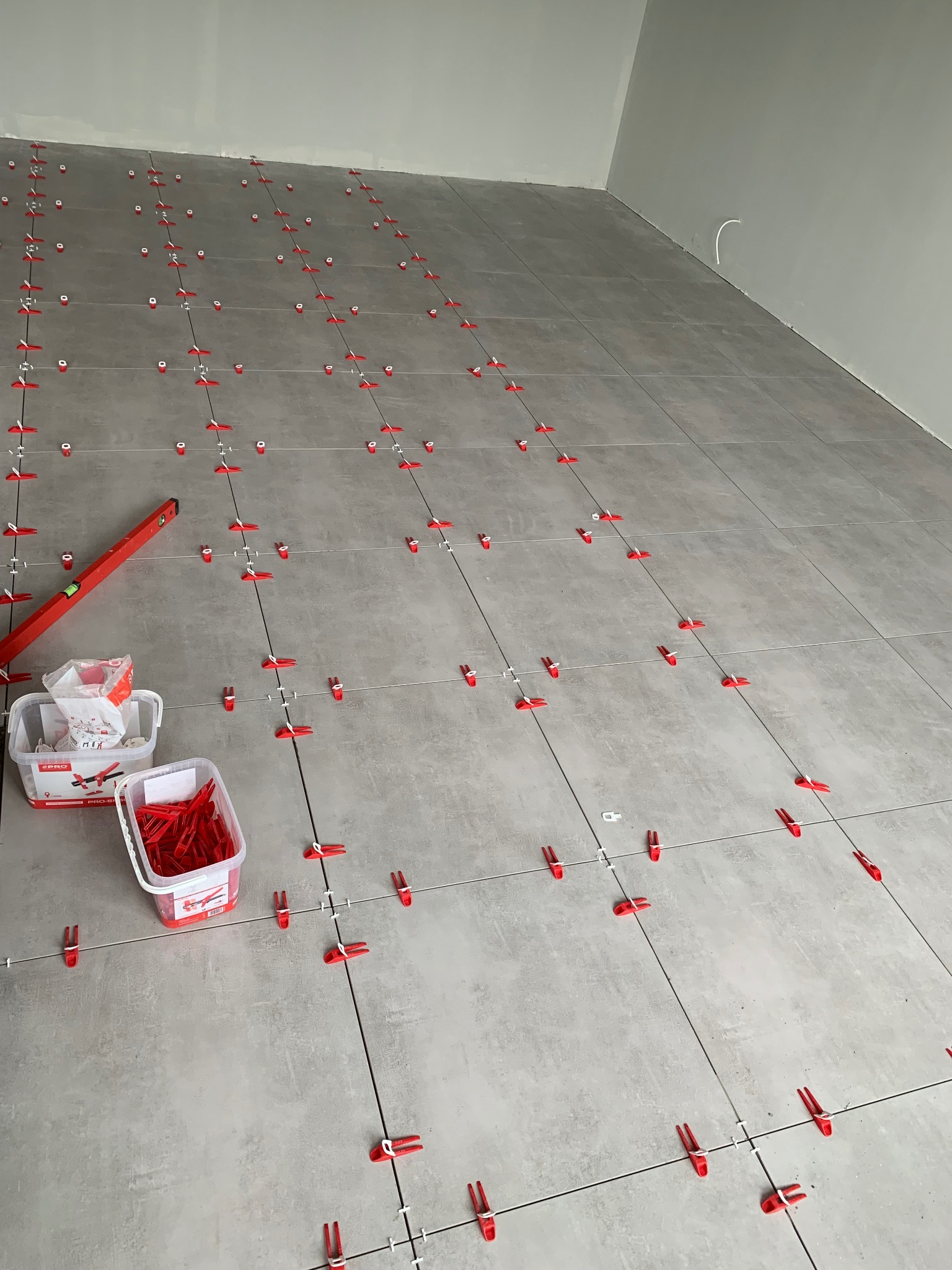 Układanie szarych płytek na podłodze z użyciem czerwonych klipsów poziomujących, widoczny poziomica i pojemniki z akcesoriami do układania płytek.