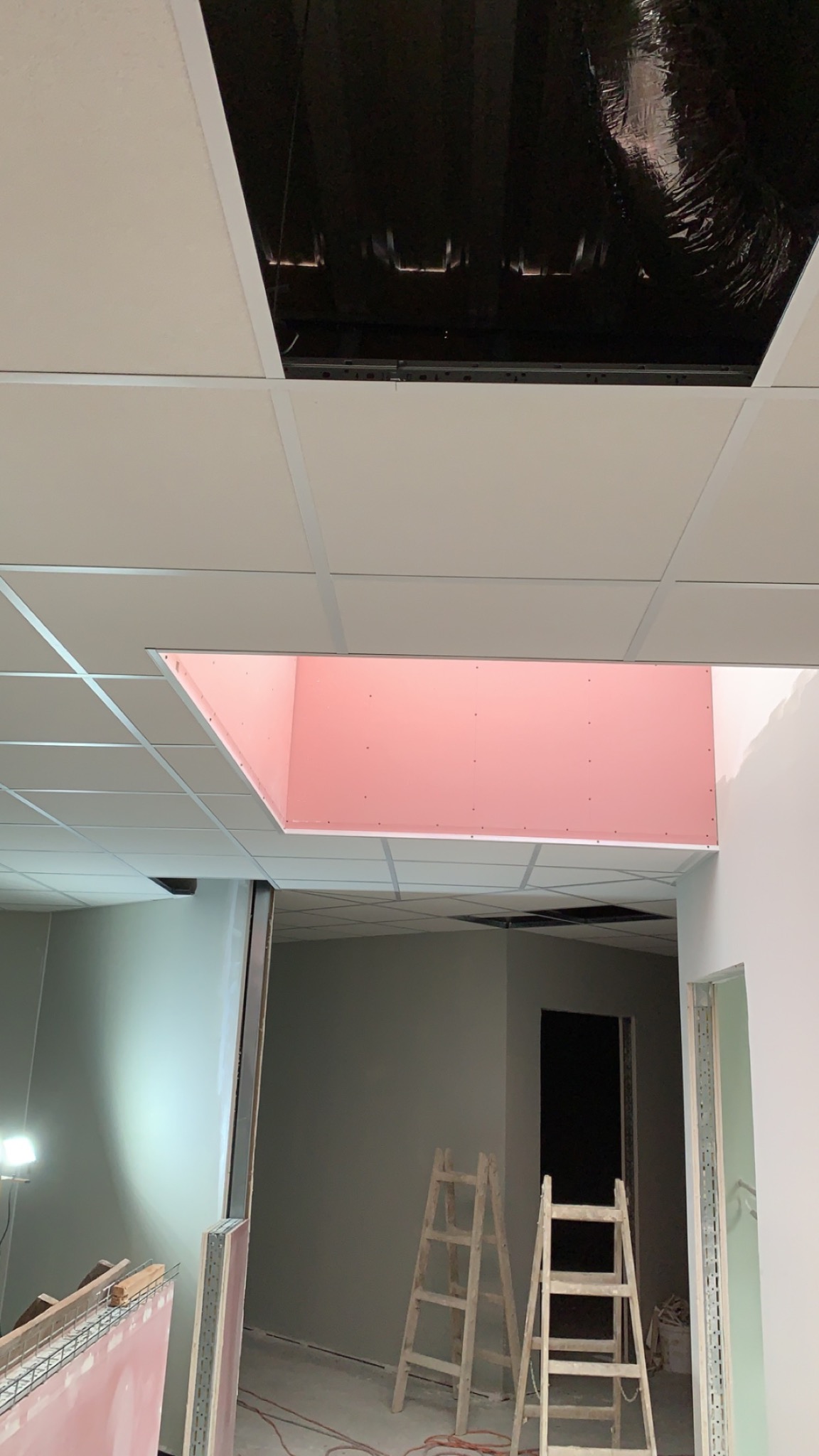 Sufit podwieszany z widoczną konstrukcją i fragmentem różowej płyty gipsowo-kartonowej w otworze, drabiny w tle, oświetlenie budowlane.