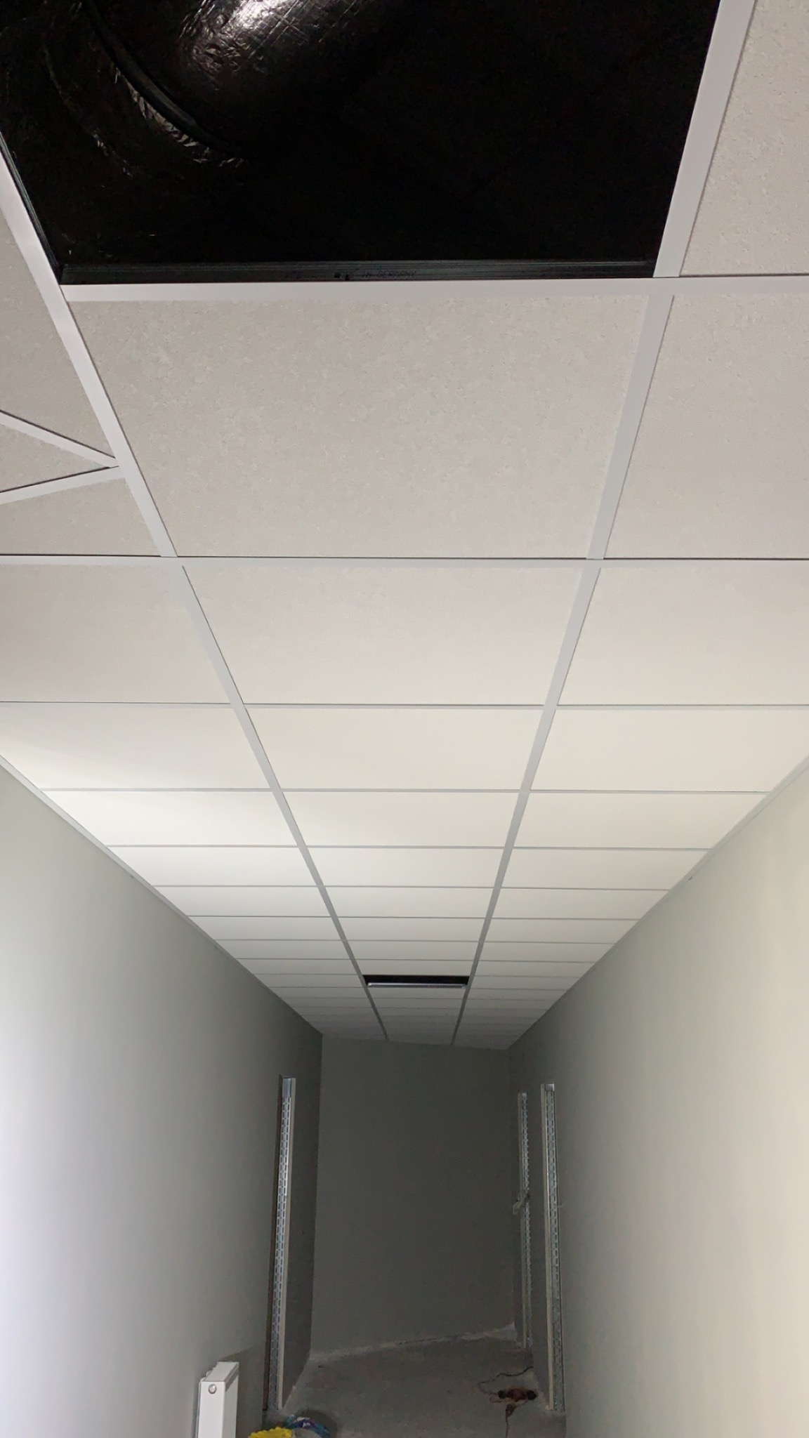 Korytarz biurowy z zamontowanym sufitem podwieszanym kasetonowym, widoczne płyty sufitowe i elementy konstrukcyjne, ściany pomalowane na szaro, fragment instalacji elektrycznej wystający z otworu...