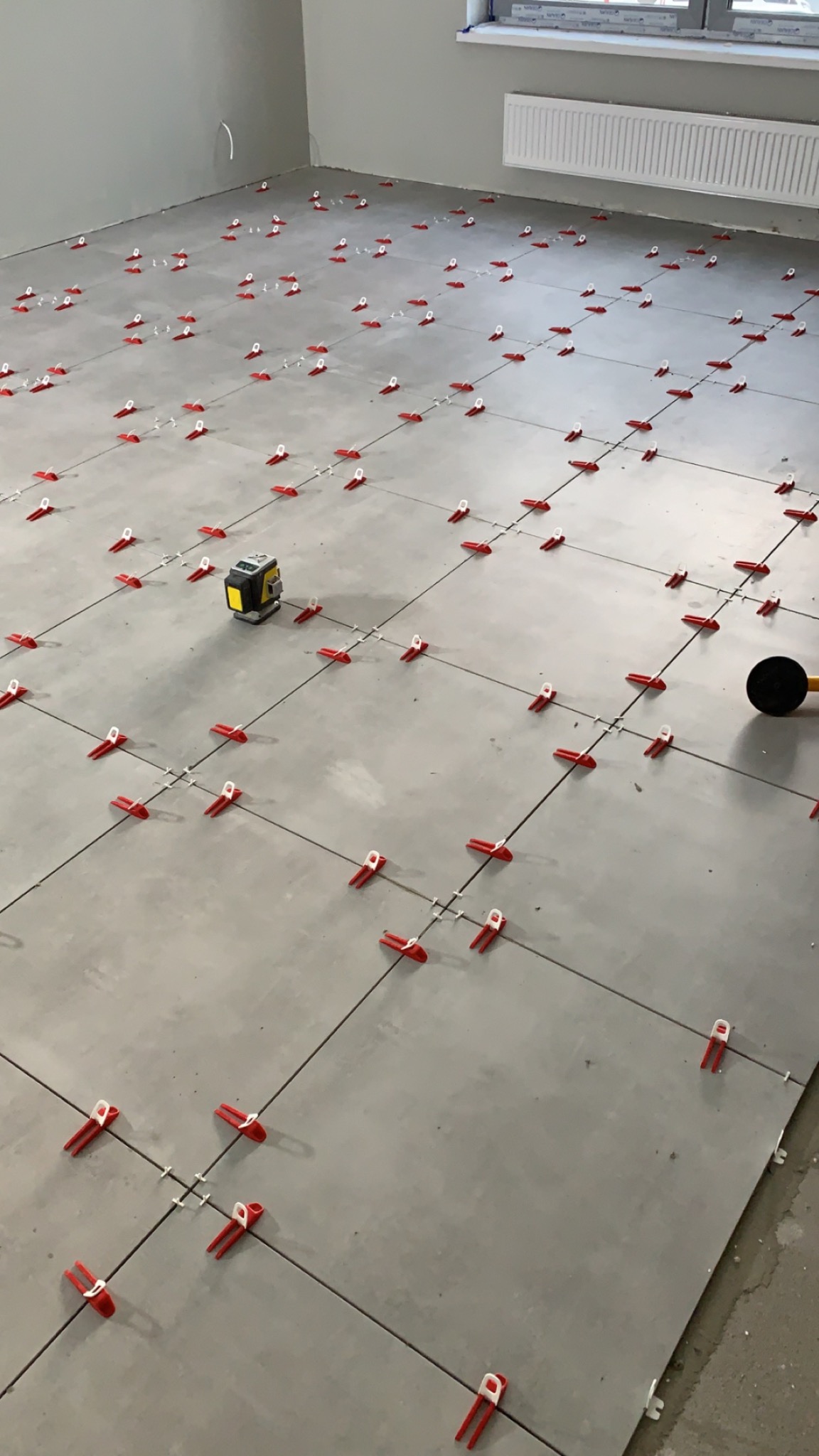 Ułożone szare płytki podłogowe z czerwonymi klipsami poziomującymi i laserem krzyżowym w rogu pomieszczenia z grzejnikiem i oknem.