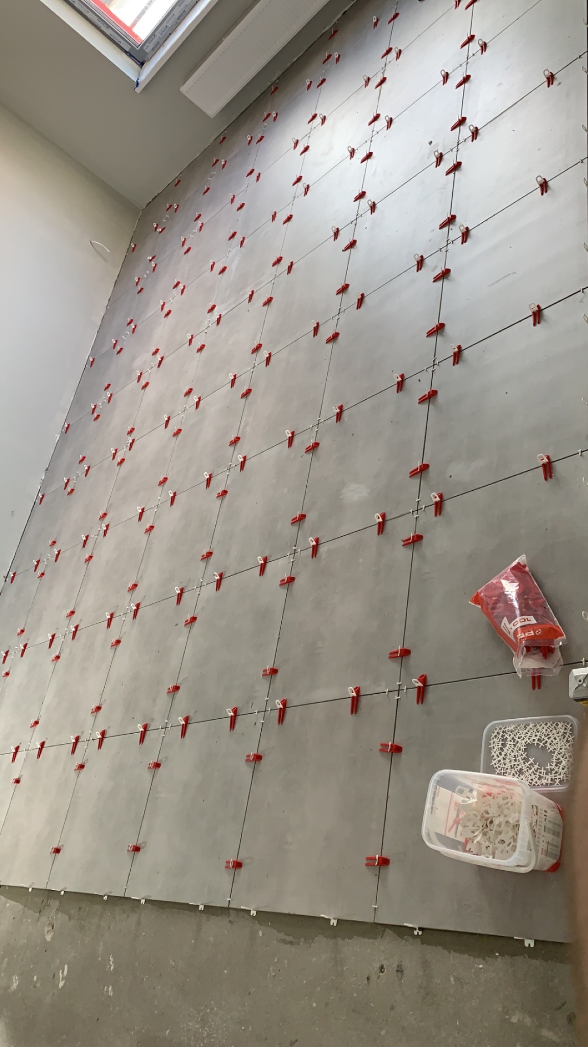 Ściana w trakcie układania szarych płytek z użyciem czerwonych klipsów poziomujących, widoczne opakowania z klipsami i klinami.