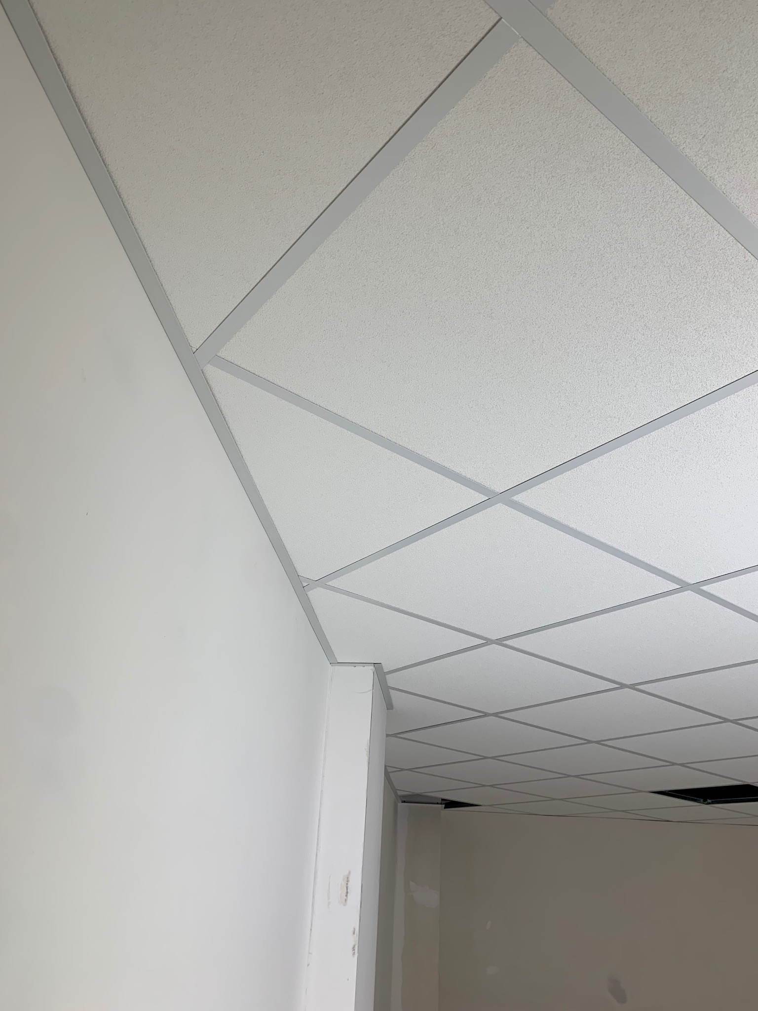 Sufit podwieszany z białych kasetonów i szarej kratownicy, widok z narożnika pomieszczenia, fragment ściany pomalowanej na biało po lewej stronie.