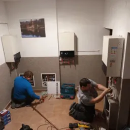 Dwóch instalatorów montuje kotły grzewcze w pomieszczeniu z beżową ścianą i obrazem zamku. Jeden instalator łączy rury miedziane, drugi pracuje przy kotle po prawej stronie. Widoczne narzędzia...