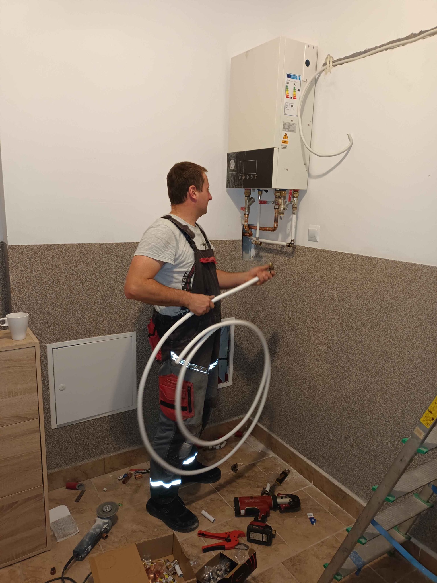 Instalator w kombinezonie roboczym montuje biały piec grzewczy na ścianie, używając rur z tworzywa sztucznego, z widocznymi narzędziami na podłodze i drabiną opartą o ścianę.