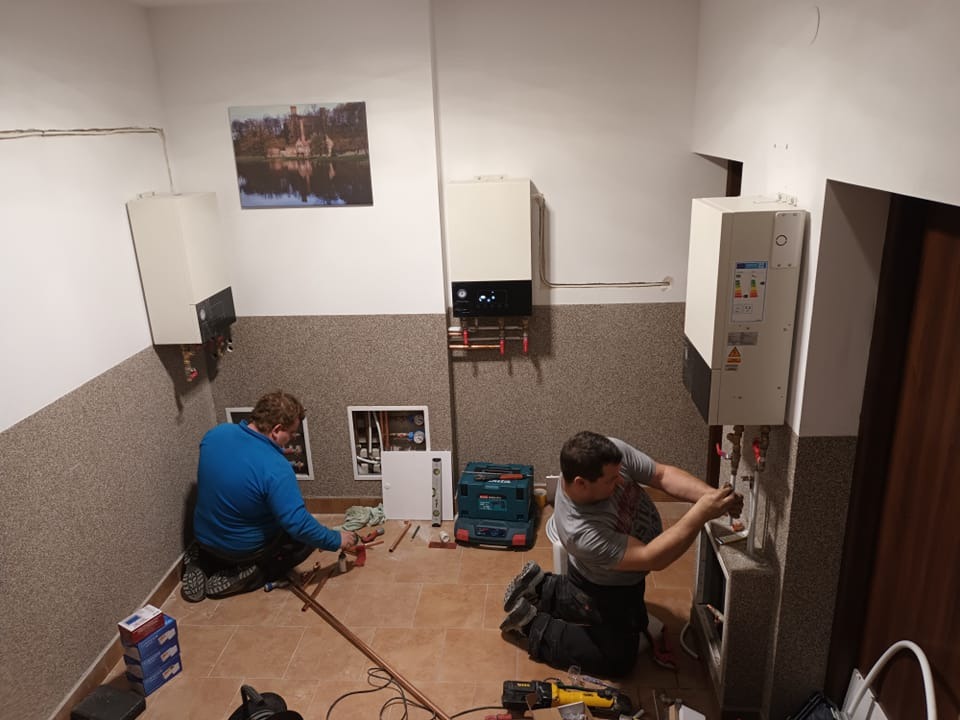 Dwóch instalatorów montuje kotły grzewcze w pomieszczeniu z beżową ścianą i obrazem zamku. Jeden instalator łączy rury miedziane, drugi pracuje przy kotle po prawej stronie. Widoczne narzędzia...