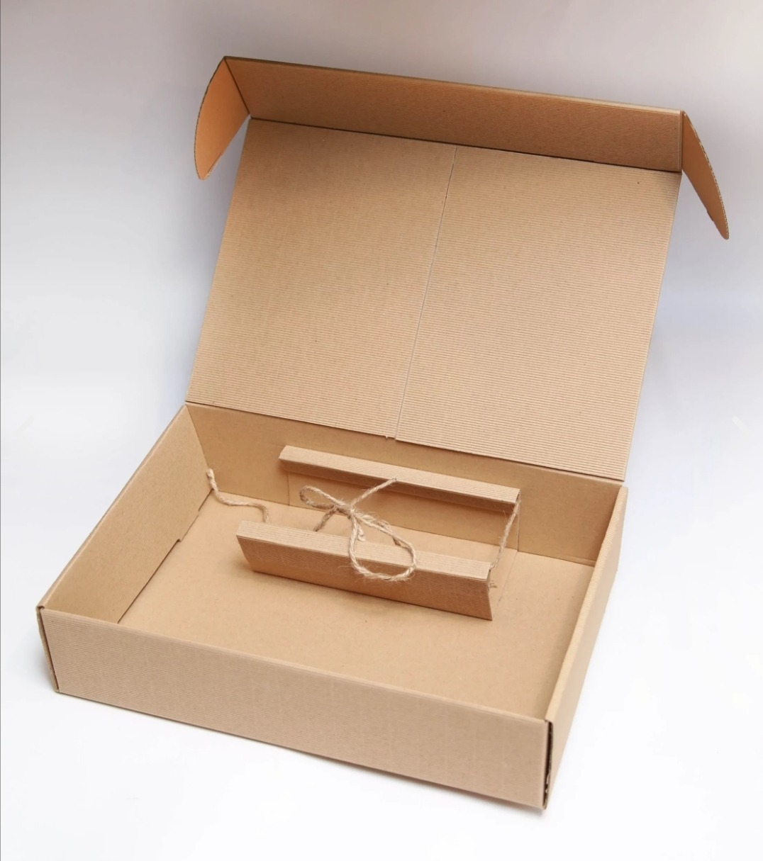 Otwarta, pusta, tekturowa skrzynka z wiekiem, w której znajduje się tekturowy element owinięty sznurkiem jutowym.