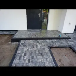 Ułożona kostka brukowa przed wejściem do domu, z czarną ramą i fragmentami ziemi. Widoczny próg i czarne drzwi wejściowe.