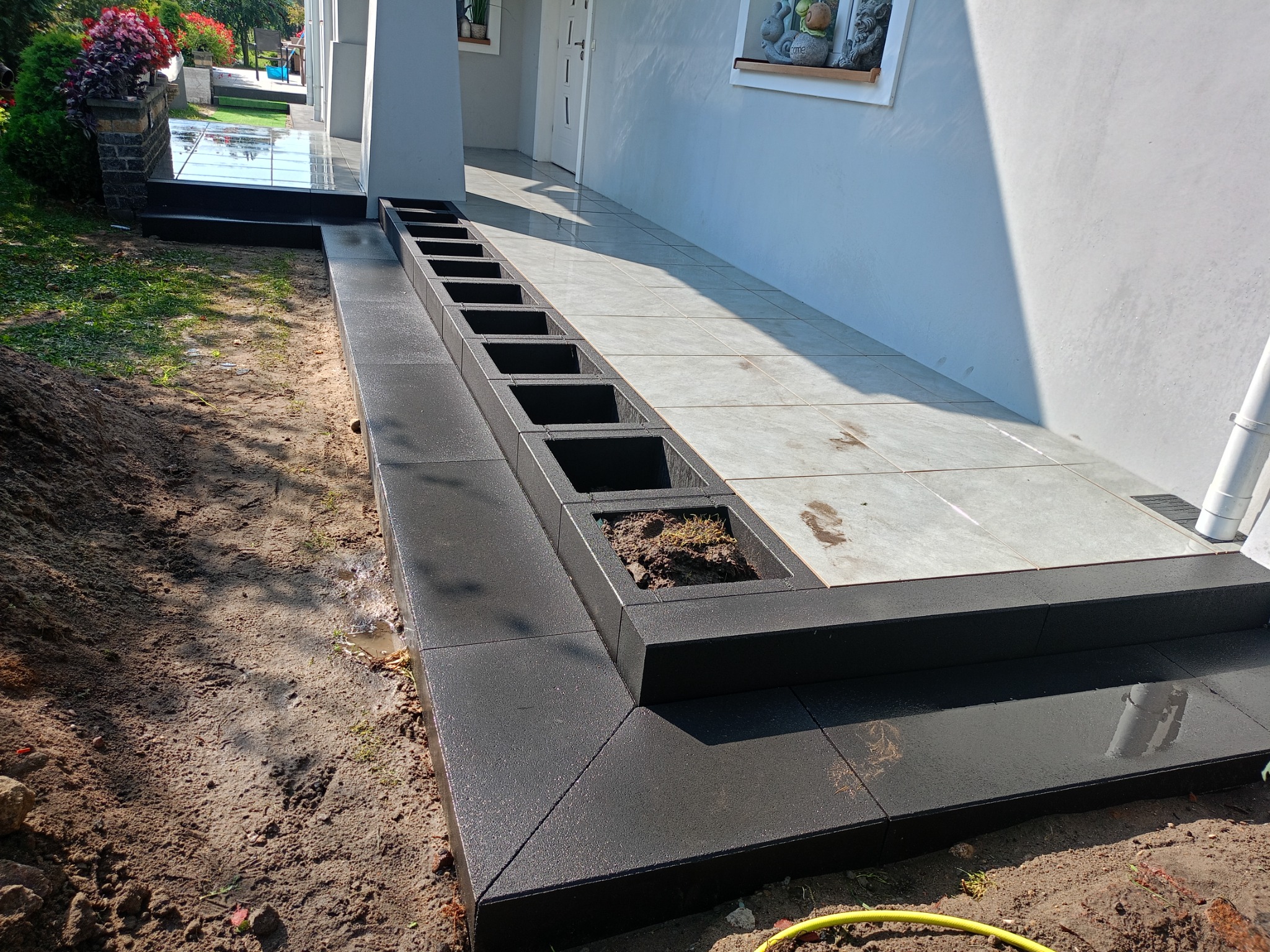 Czarne, matowe stopnie z kostki brukowej z wbudowanymi donicami, prowadzące do jasnego tarasu przy domu.