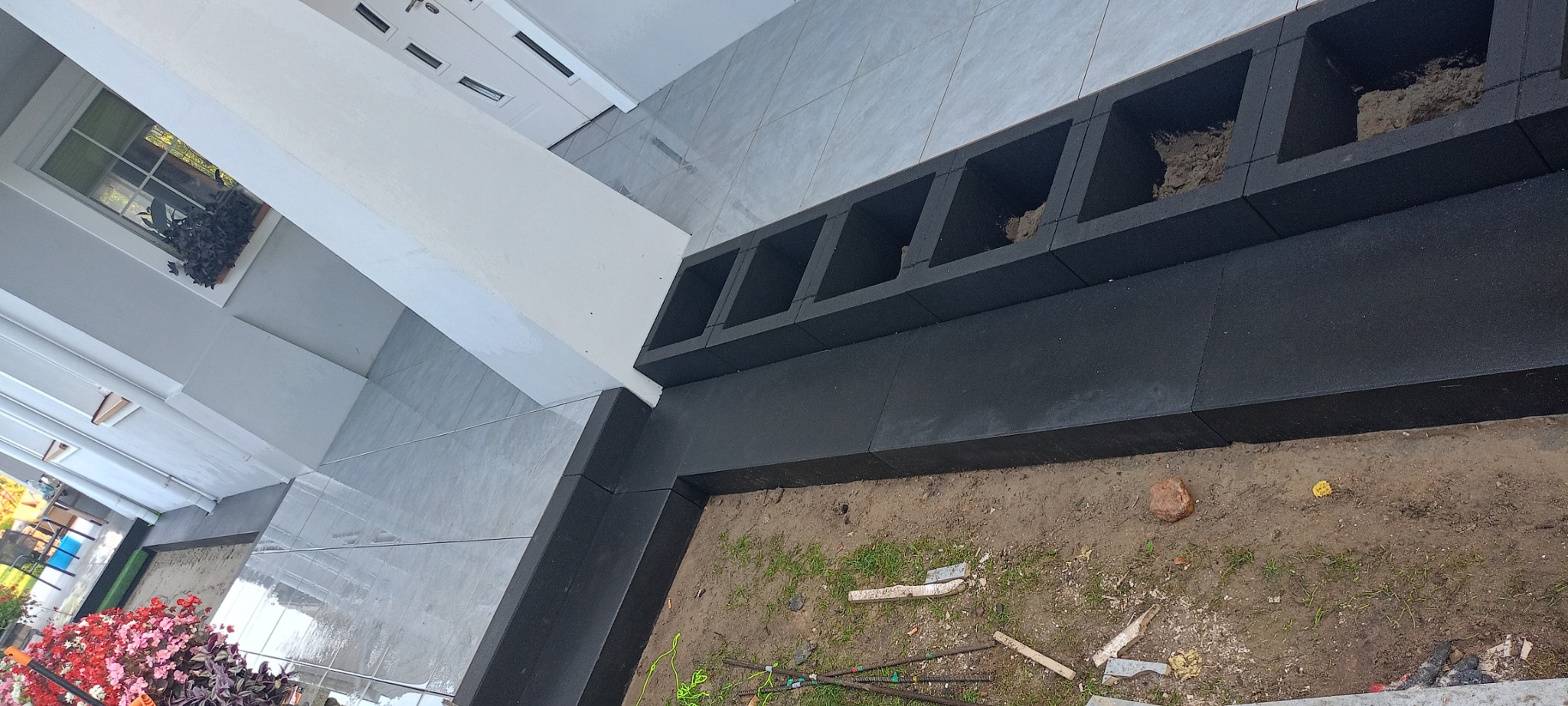 Czarne, modułowe obrzeże z pustymi przestrzeniami, prawdopodobnie na rośliny, oddzielające płytki chodnikowe od rabaty ziemnej z widocznymi fragmentami drewna i prętów zbrojeniowych.