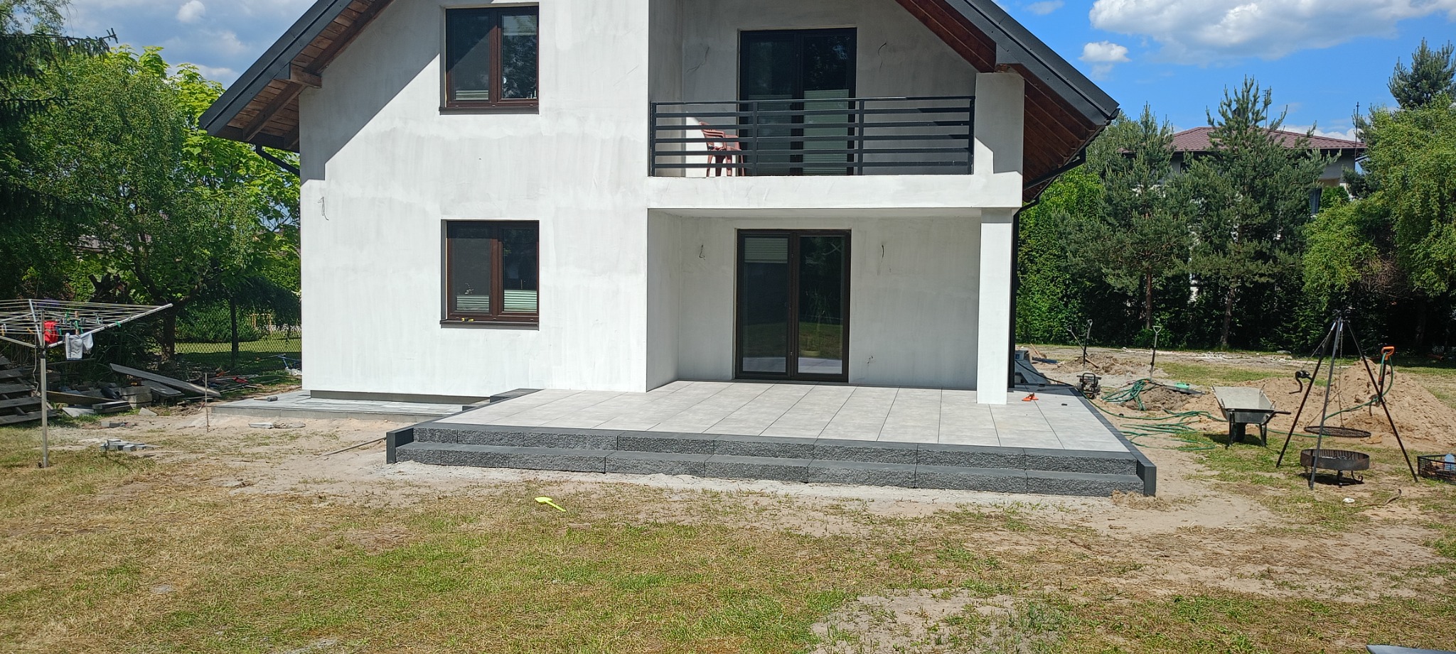 Nowo wyłożony taras z szarych płytek z dwustopniowymi schodami z ciemniejszej kostki, na tle białego, dwupiętrowego domu z balkonem i zielenią ogrodową.