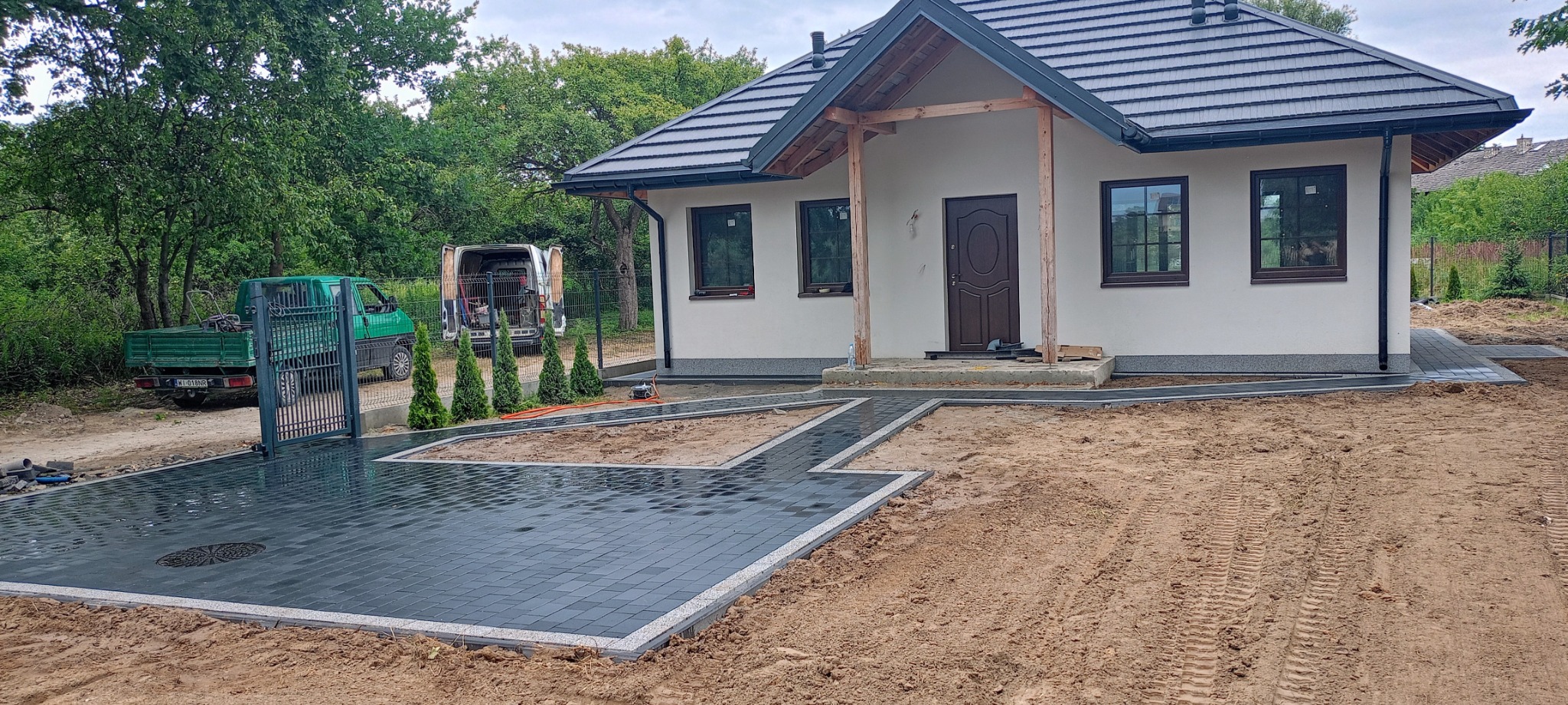 Ułożona, ciemnoszara kostka brukowa z jasnoszarym obramowaniem prowadzi do domu z białymi ścianami i ciemnym dachem; w tle zaparkowane dwa samochody dostawcze i zieleń.