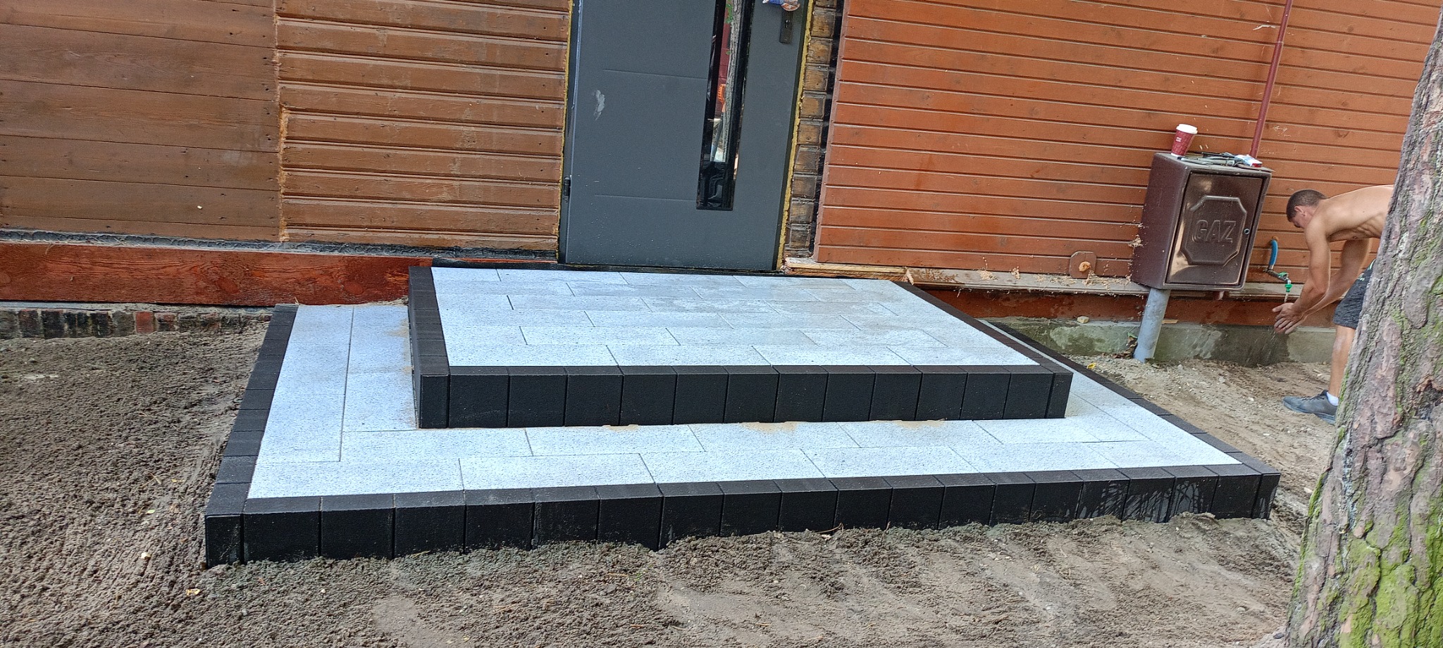 Nowe dwustopniowe schody z jasnoszarej kostki brukowej z czarnym obramowaniem, prowadzące do drewnianego domu w Otwocku. W tle widoczny mężczyzna myjący ręce przy skrzynce gazowej.