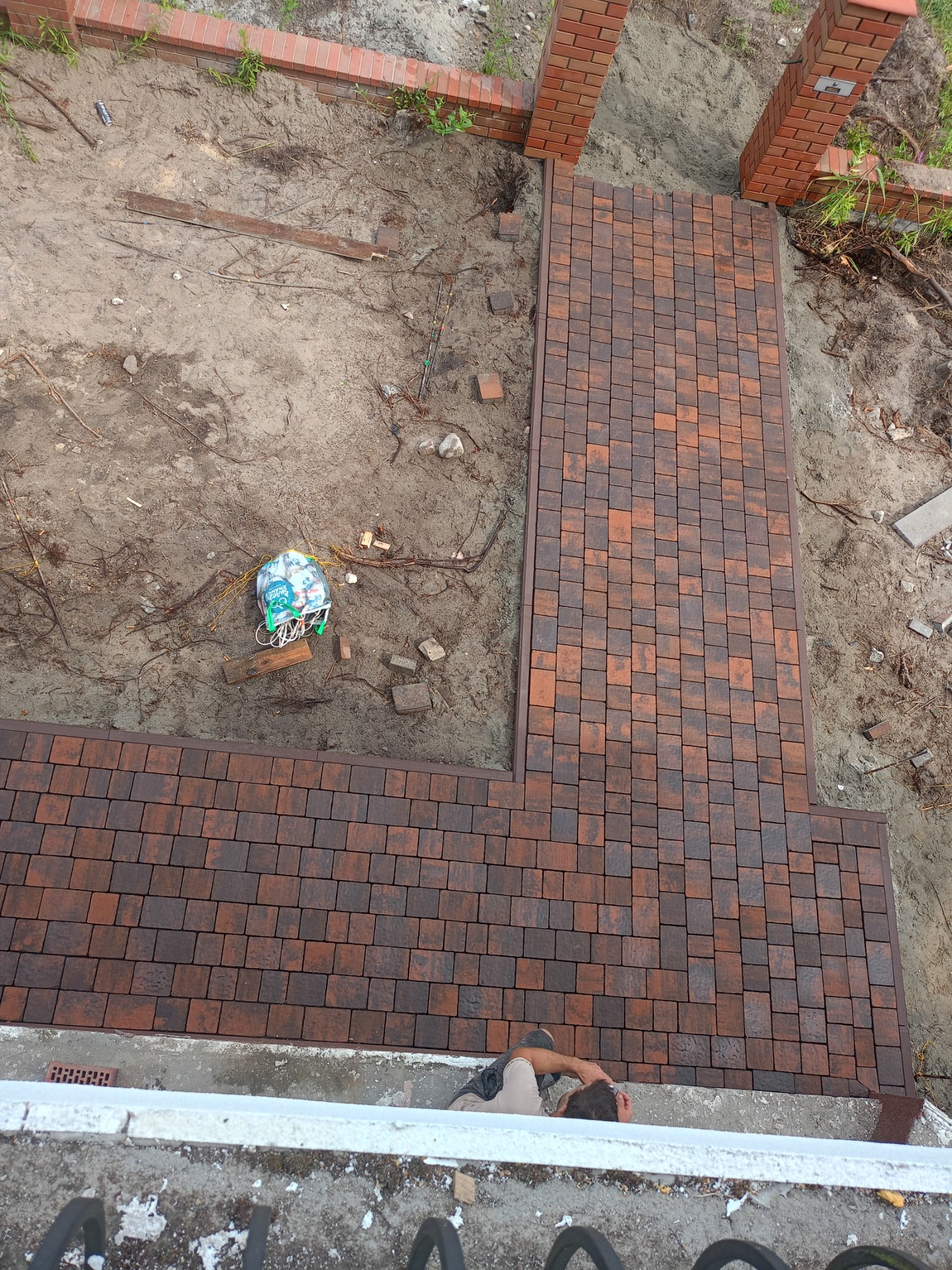 Widok z góry na częściowo ułożoną ścieżkę z kostki brukowej w odcieniach brązu i czerwieni, z widocznymi elementami przygotowania podłoża, takimi jak piasek i resztki materiałów budowlanych...