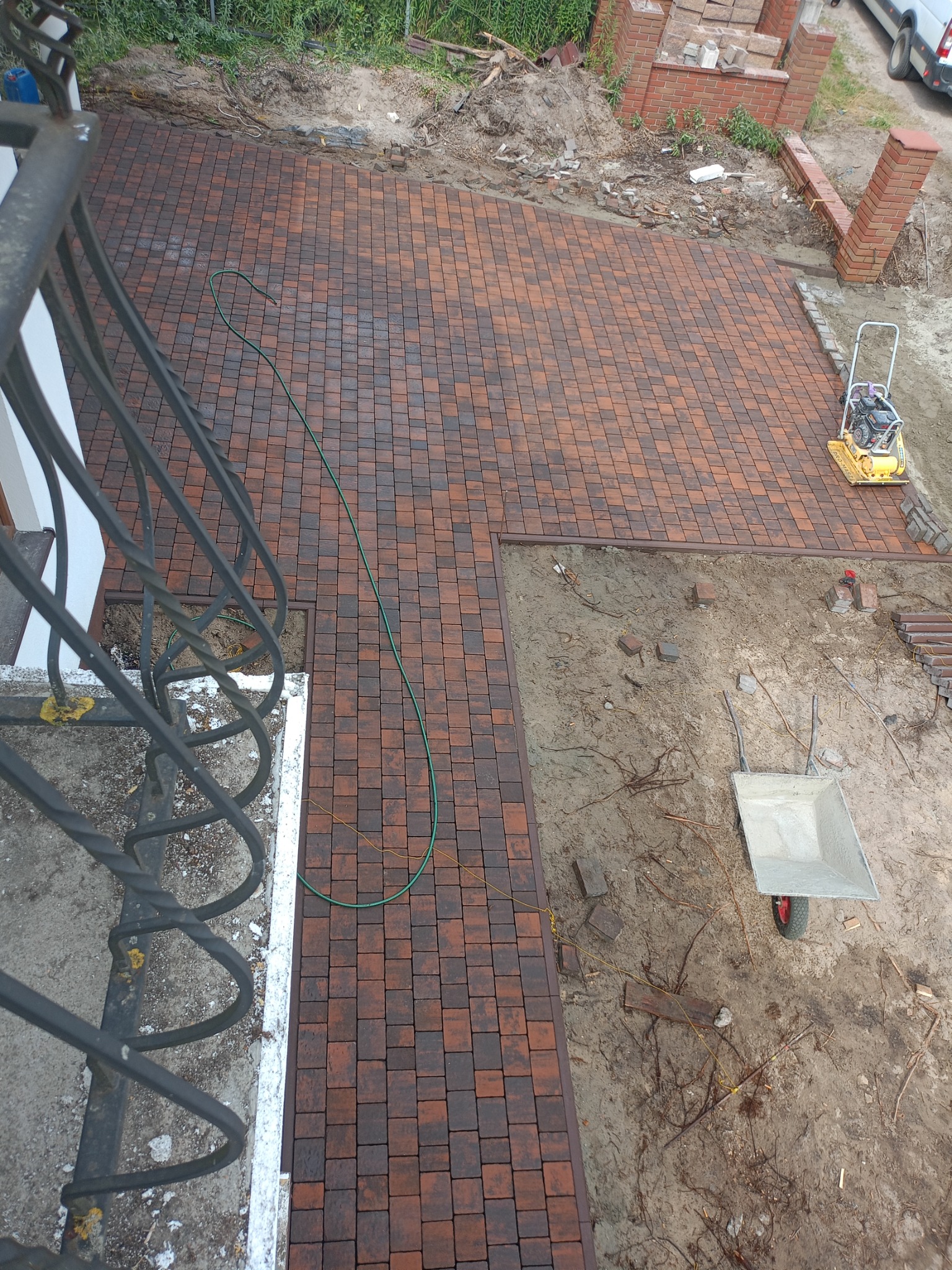 Widok z góry na częściowo ułożoną, brązowo-czerwoną kostkę brukową, widoczna zagęszczarka, taczka i elementy muru z cegły.