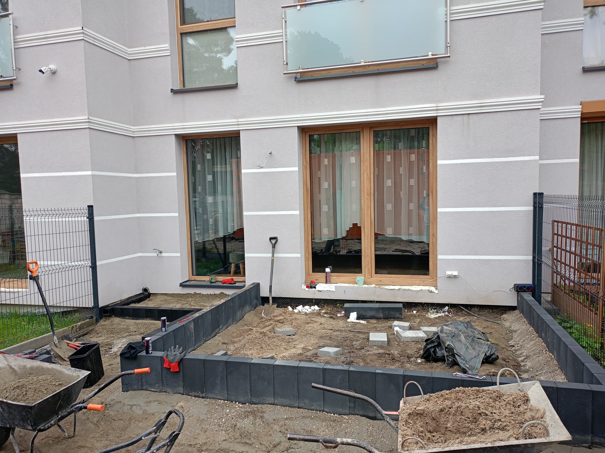 Przygotowanie podłoża pod taras z kostki brukowej: czarne obrzeża, piasek, narzędzia i taczki w ogrodzie przed budynkiem z oknami.