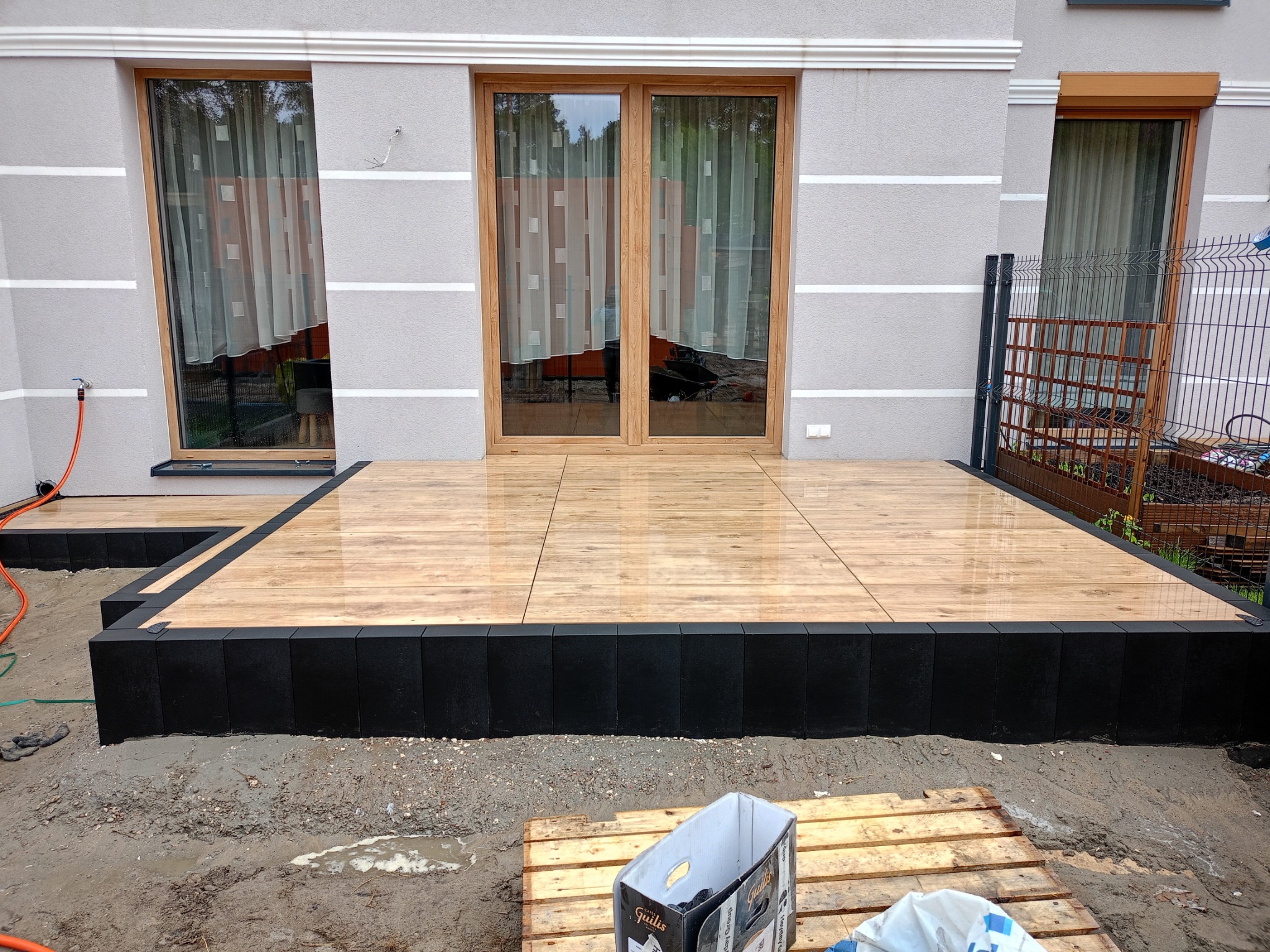 Nowoczesny taras z drewnopodobnych płytek ceramicznych obramowany czarnymi obrzeżami na tle szarej elewacji domu z drewnianą stolarką okienną, widoczne ślady prac ziemnych i paleta z pudełkiem po...