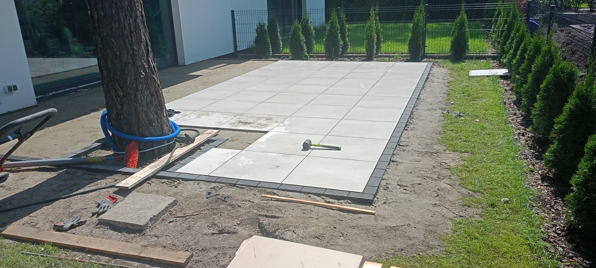 Montaż jasnych, kwadratowych płyt tarasowych z ciemnoszarą obwódką wokół drzewa, widoczne narzędzia i rękawice robocze