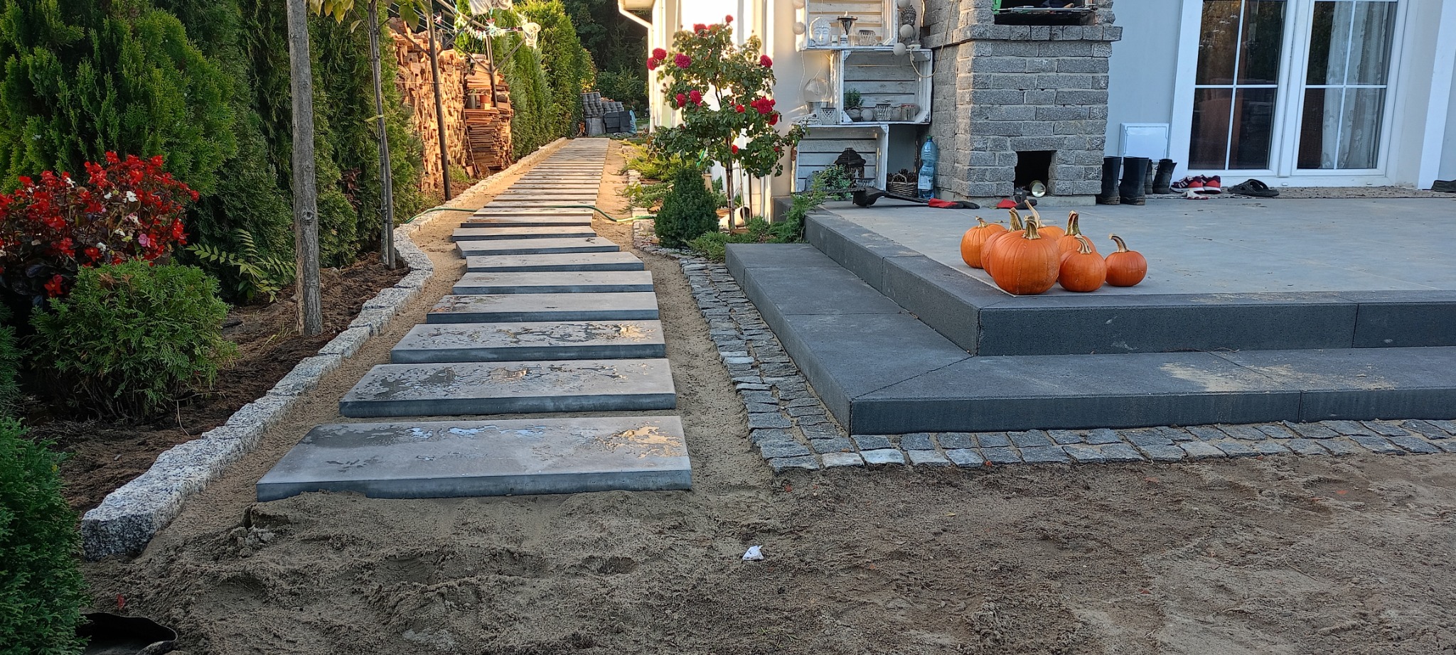 Nowoczesna ścieżka z płyt betonowych prowadząca przez ogród, obrzeża z kostki granitowej, schody z ciemnych płyt z dyniami na stopniach, w tle dom z elementami z szarej cegły.