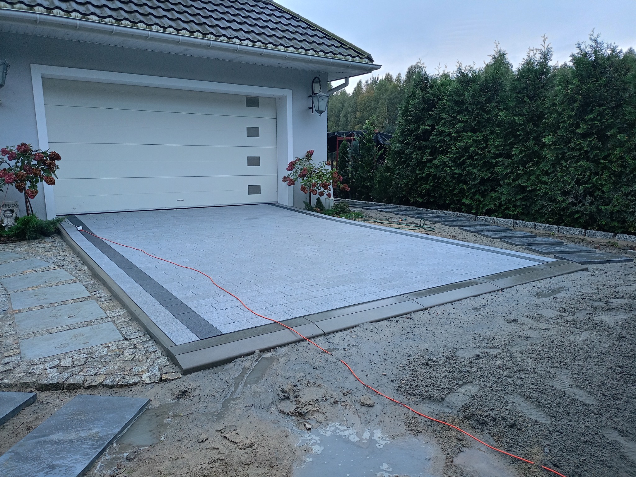 Nowa nawierzchnia z szarej kostki brukowej przed garażem, obrzeża z ciemniejszej kostki, widoczny kabel na piasku.