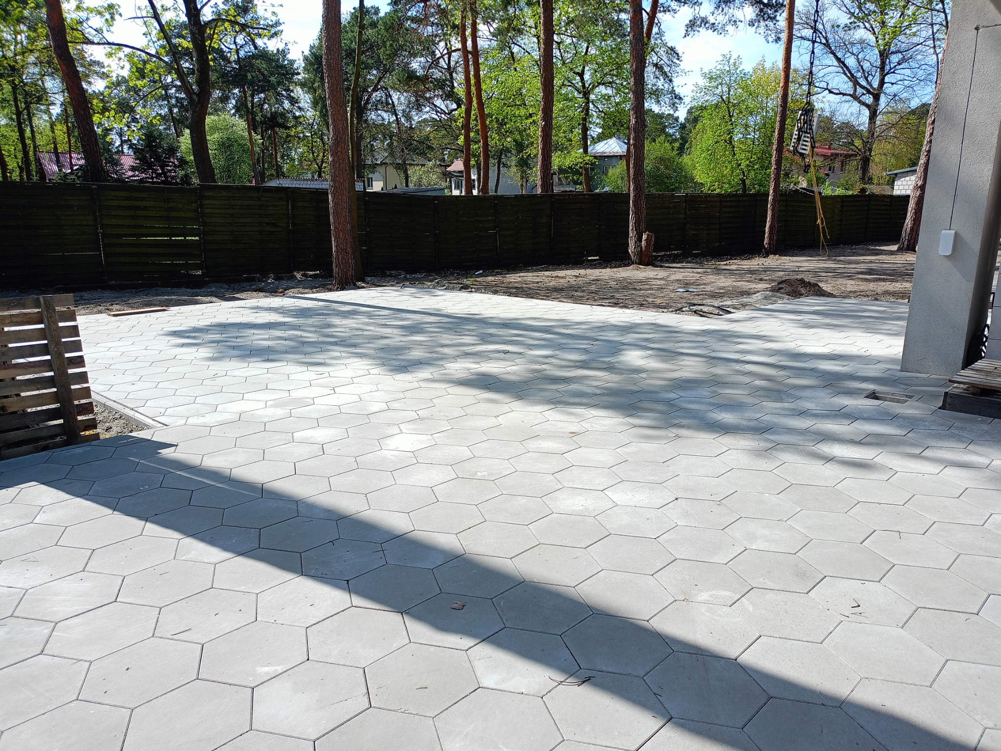 Ułożona, heksagonalna kostka brukowa w odcieniach szarości na posesji z drzewami i drewnianym ogrodzeniem w tle, widok z góry.