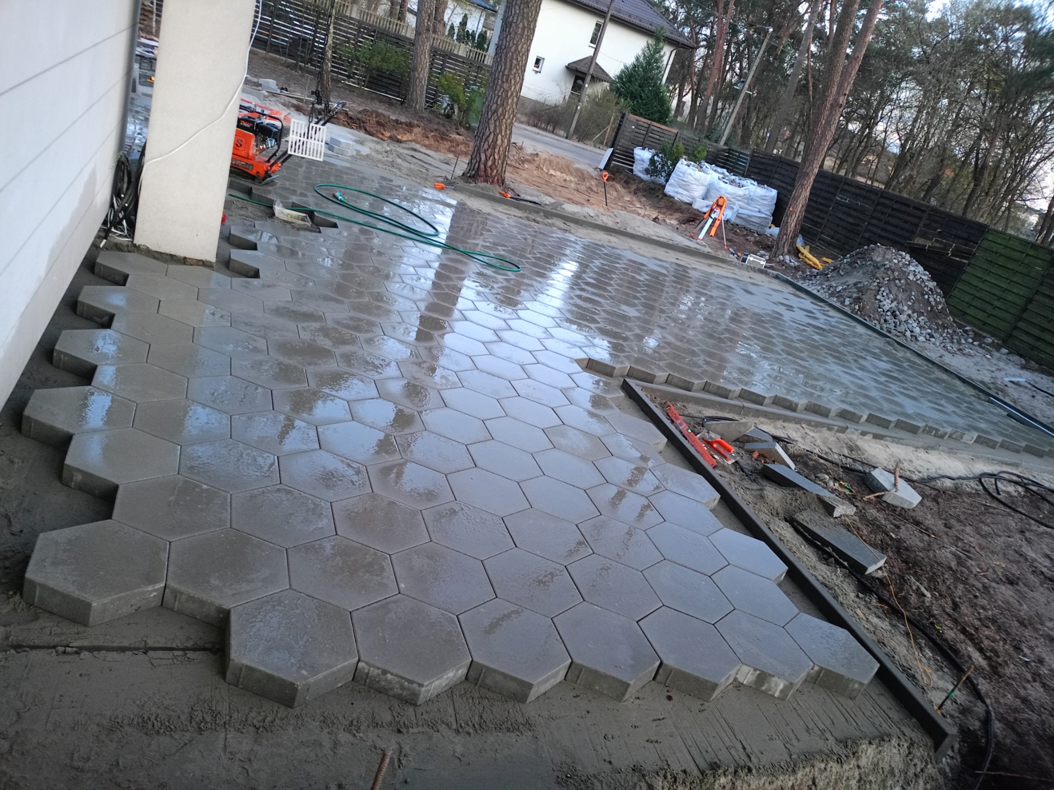 Układanie heksagonalnej kostki brukowej w ogrodzie, widoczny zagęszczarka, przygotowane podsypki i narzędzia. Częściowo ułożona nawierzchnia.