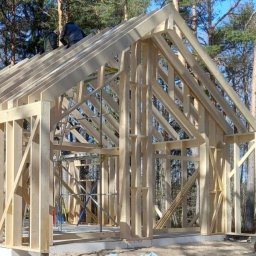 woodenvill - Szkielet drewniany domu w trakcie budowy, widoczne elementy konstrukcyjne i dwóch pracowników na dachu, las w tle, słoneczny dzień.