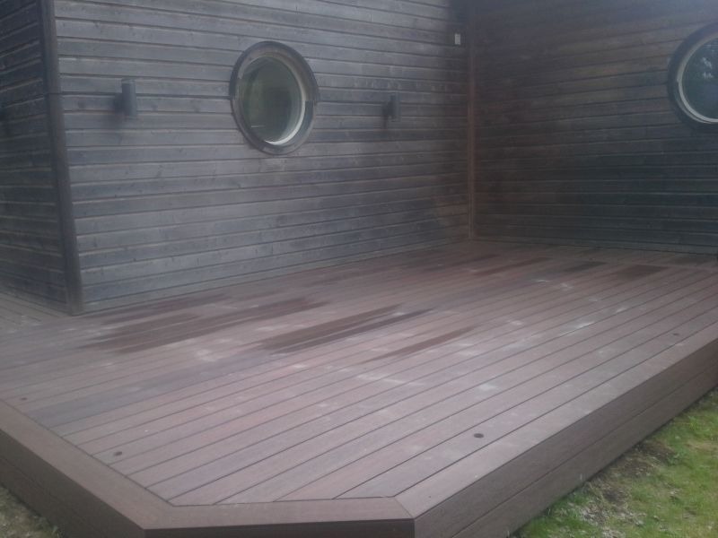 Nowoczesny taras kompozytowy przylegający do budynku z oknami w kształcie bulaja. Deski ułożone poziomo, widoczne mocowania. Tło stanowi elewacja budynku.